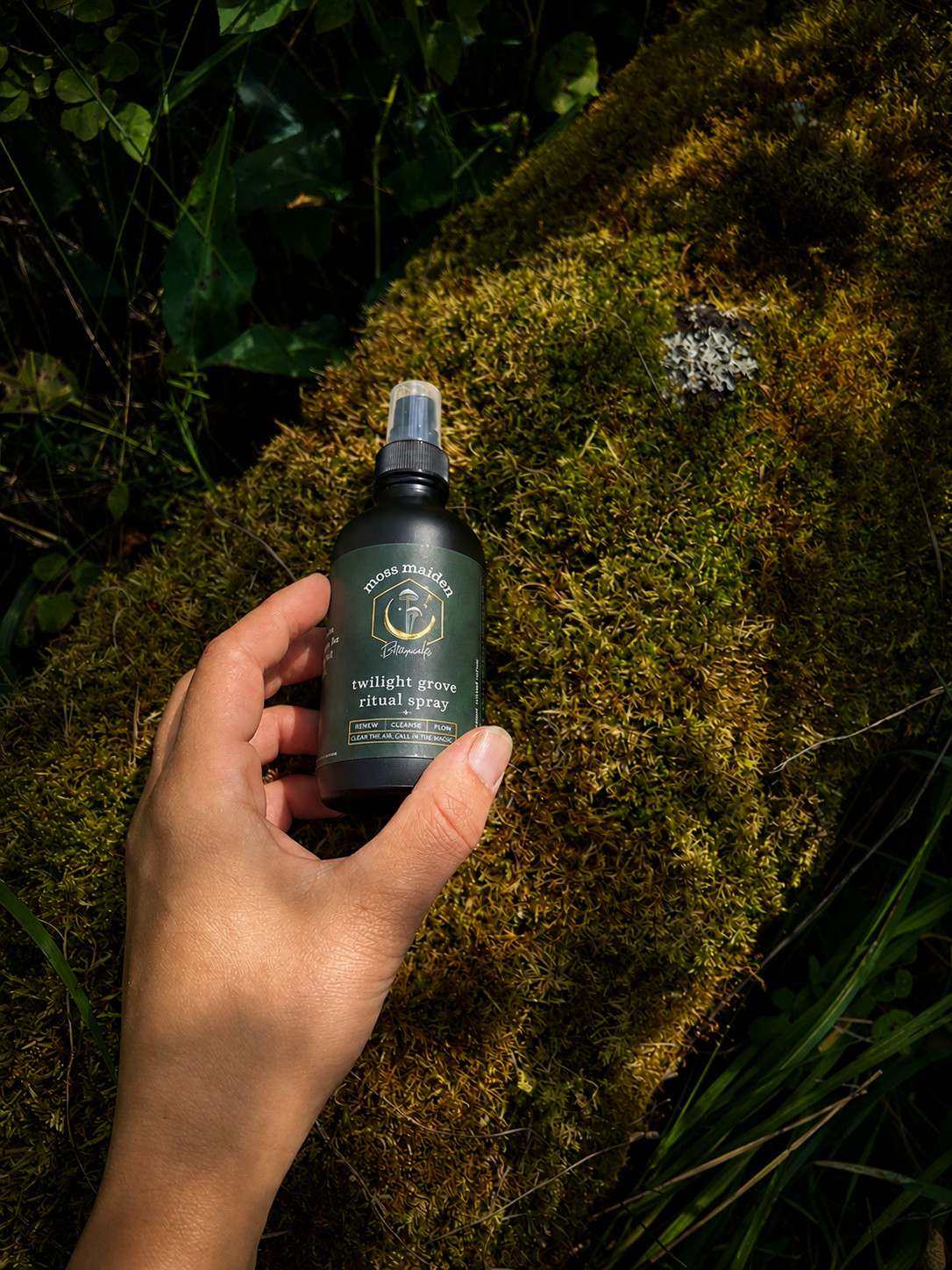 A hand holding a black bottle labeled 'Moss Maiden Twilight Grove Ritual Spray' over moss on the ground in a natural outdoor setting.