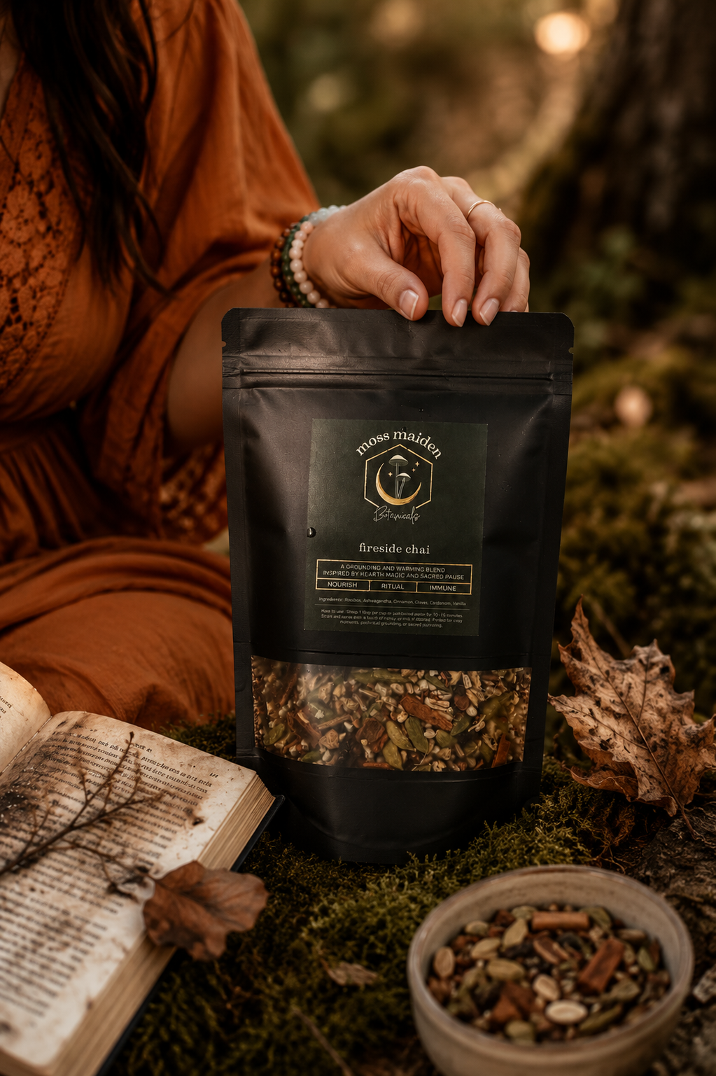 A person holding a black package labeled 'Moss Maiden Fire Side Chai' in a forest setting, with an open book and a small bowl of loose tea spices on the mossy ground nearby.