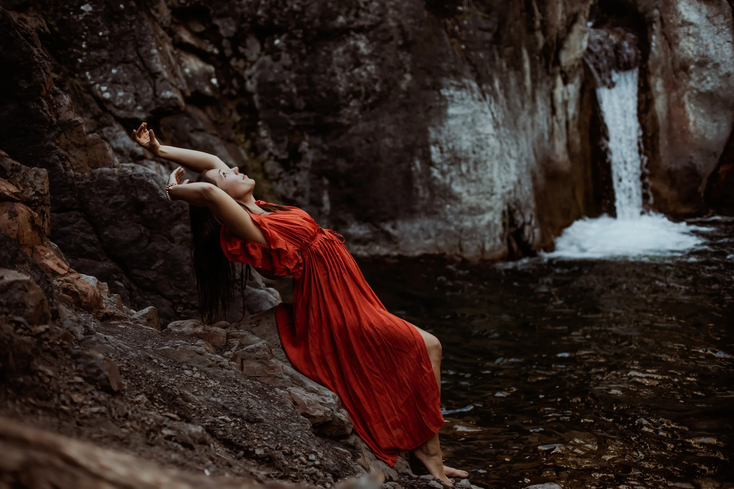 A woman in a red dress leaning against rocks near a waterfall in a rocky outdoor setting.