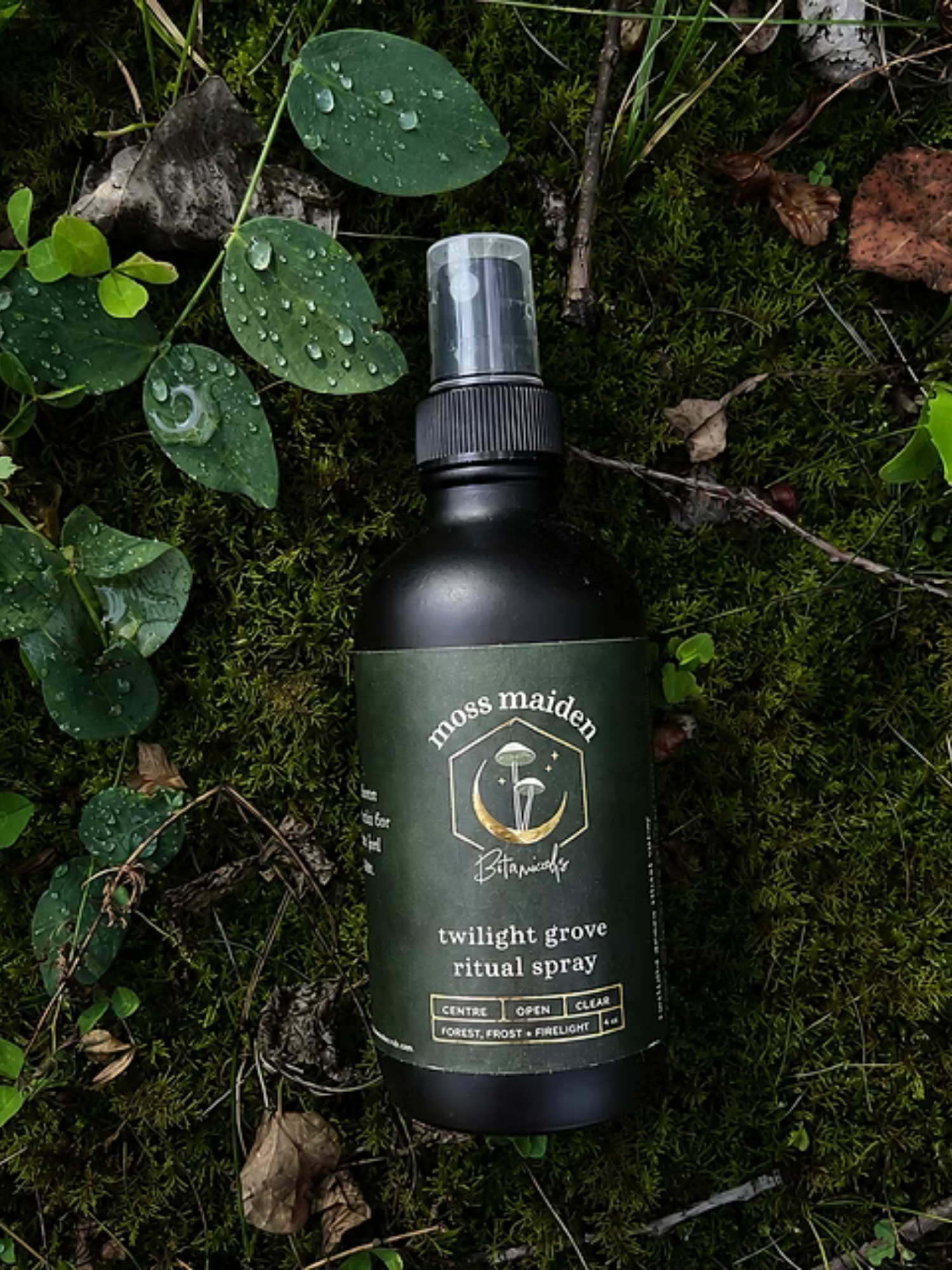A bottle of Twilight Grove ritual spray labeled 'moss maiden' lying on moss and leaves in a forest setting.