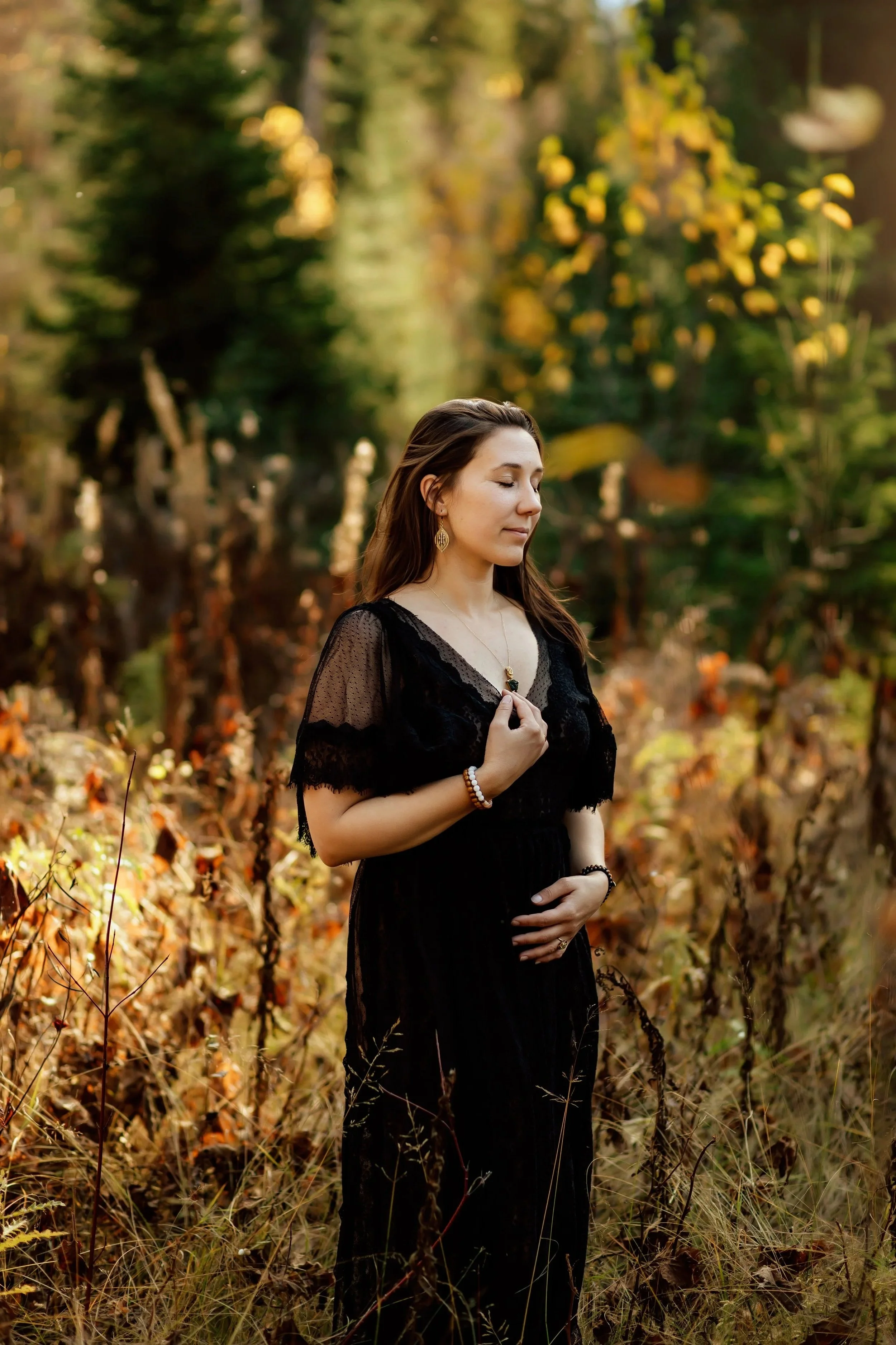 A woman with long brown hair, wearing a black lace dress and jewelry, standing outdoors in a forest with autumn leaves and trees in the background.