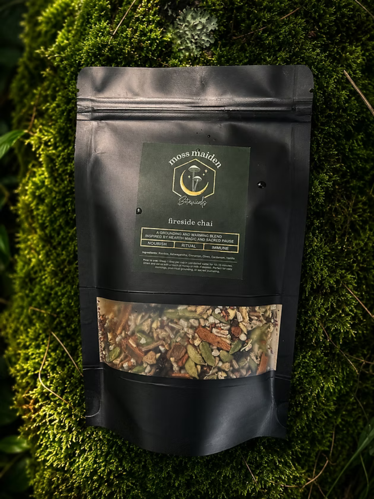 A black resealable bag labeled 'moss maiden botanica' with a logo of a mushroom and moon. The bag contains a herbal blend called 'fireside chai' and is placed on a bed of moss and greenery.