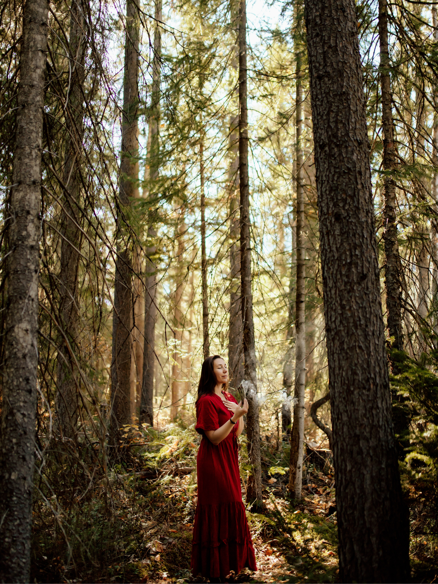 A woman in a long red dress standing in a forest among tall trees, with her eyes closed and hands over her chest, holding a bunch of sticks or twigs.