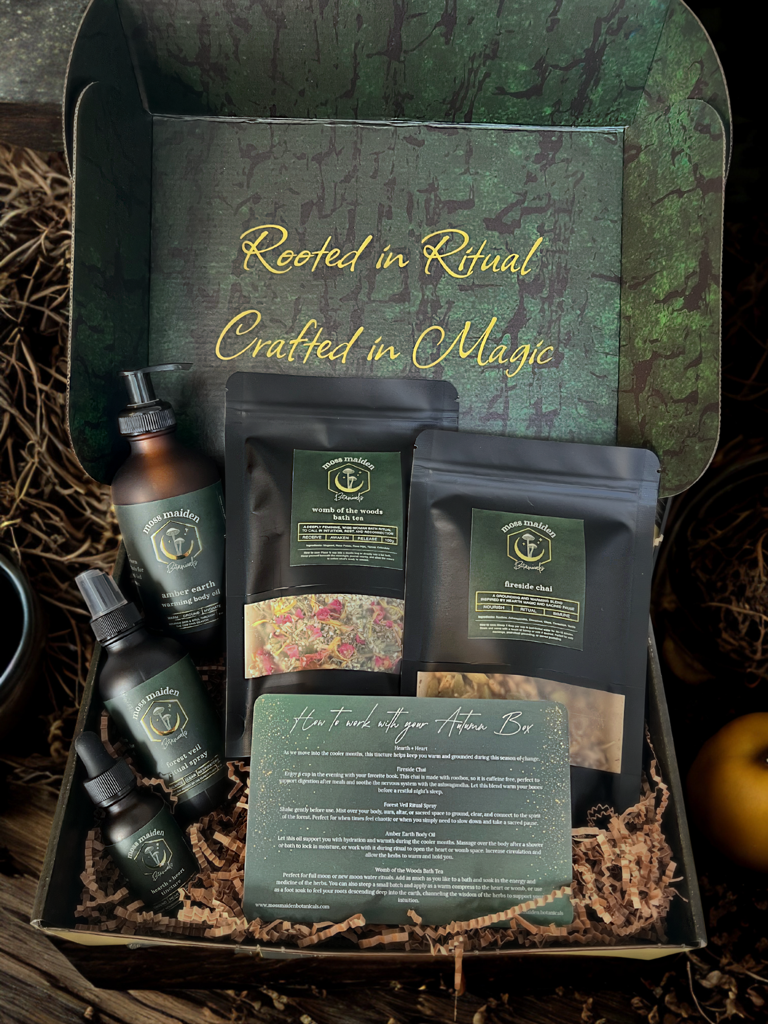 An open gift box with a nature-themed interior, displaying various herbal products including body oil, bath tea, and herbal sprays. The background of the box features a green and brown bark pattern with the phrases "Rooted in Ritual" and "Crafted in Magic" written inside.