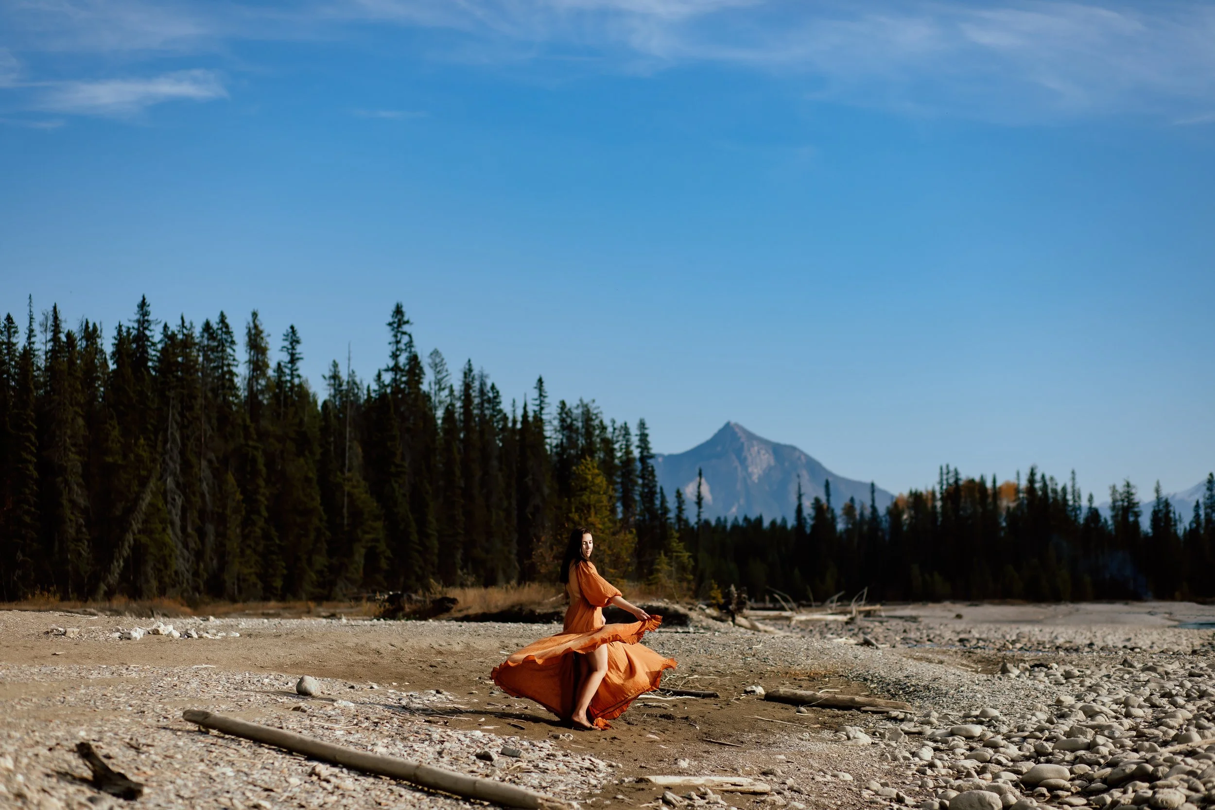 A woman in a flowing brown dress sitting on a sandy, rocky shoreline with forested mountains and a blue sky in the background.