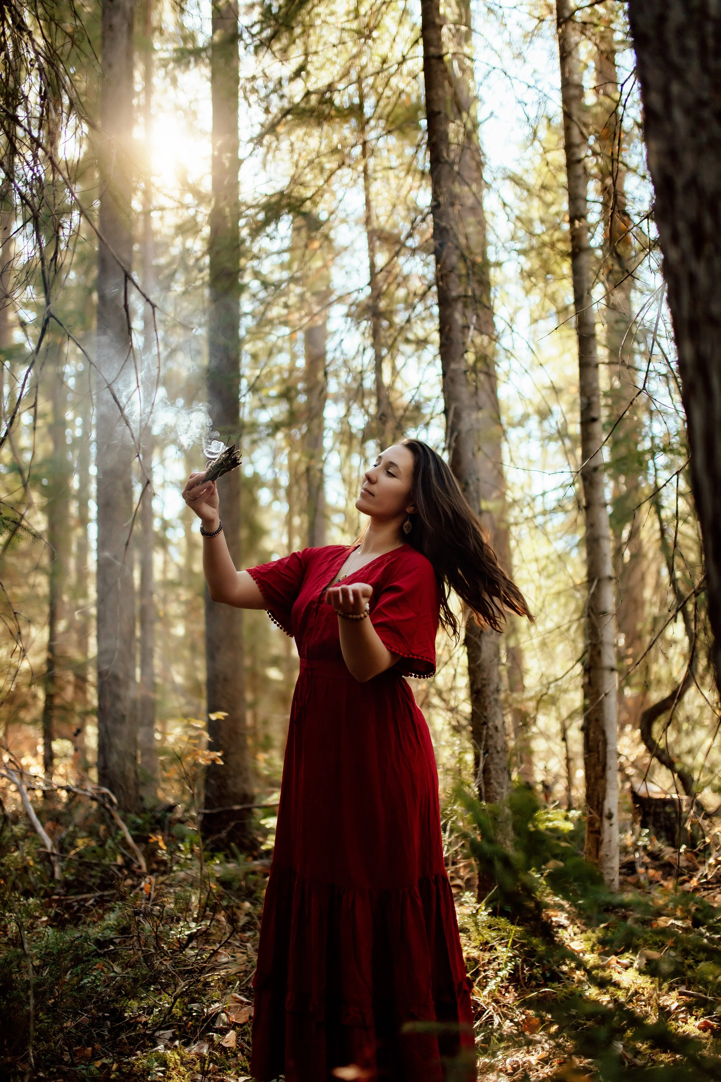 A woman with long dark hair and a red dress standing in a forest holding a lit bundle of sticks with smoke rising from it, sunlight filtering through trees.