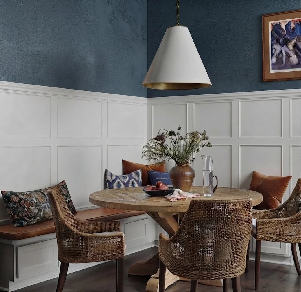 A cozy dining area with a round wooden table, four woven rattan chairs, and a built-in white bench with colorful cushions. A large vase with flowers and a pitcher of water sit on the table. The background features dark blue walls with white wainscoting and a framed artwork hanging on the wall. A white pendant light hangs above the table.