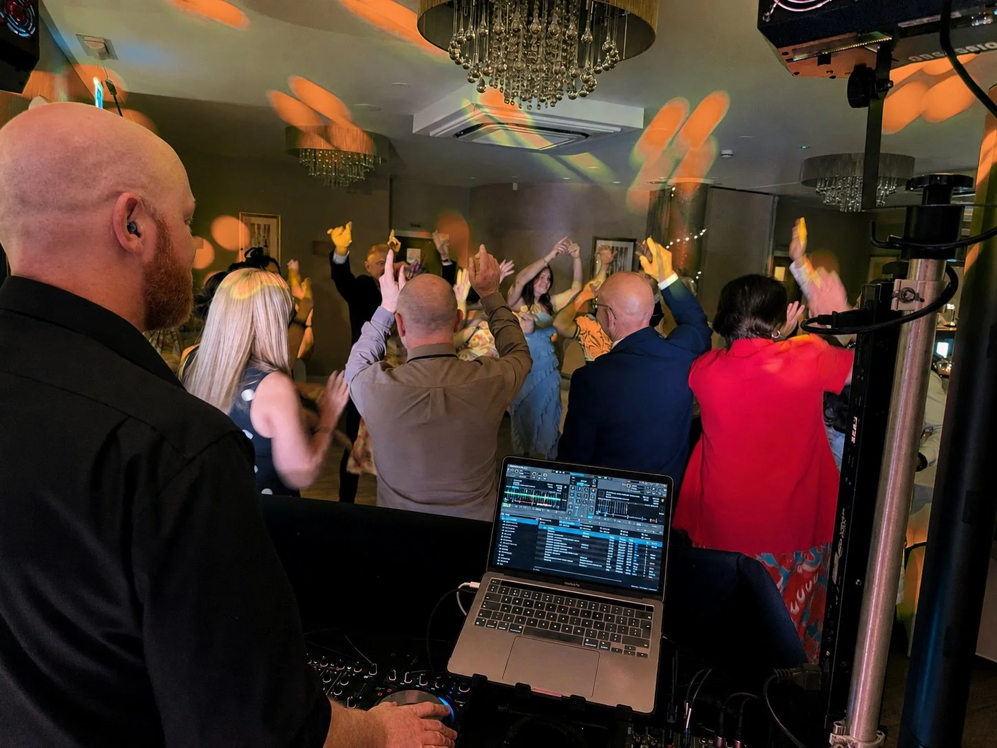People dancing and celebrating at a party with a DJ playing music in the foreground, surrounded by colorful lighting and chandeliers.