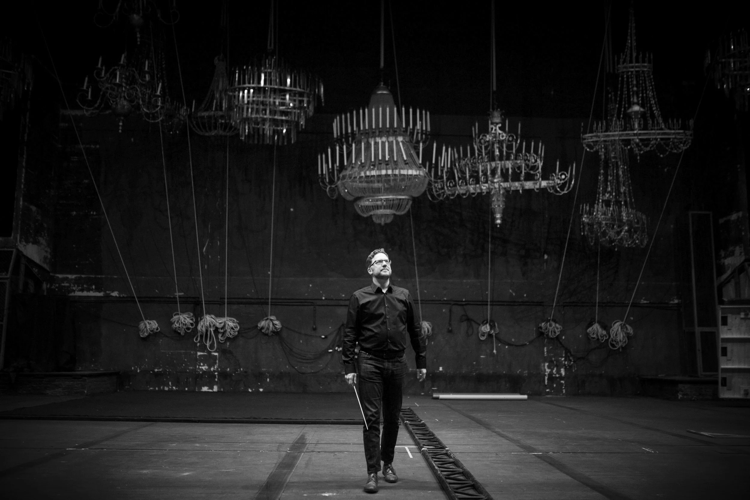 Black-and-white photo of Benjamin Bayl on theater stage beneath numerous ornate chandeliers