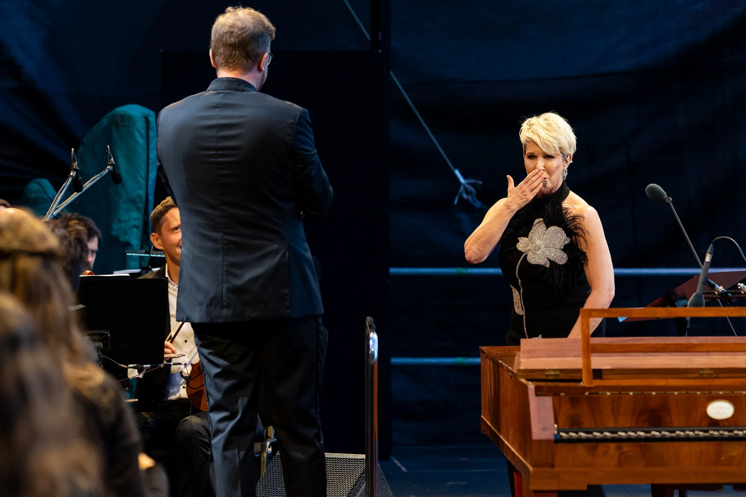 Blonde female performer in a black floral dress blows a kiss onstage beside a conductor, musicians, microphones, and a piano.