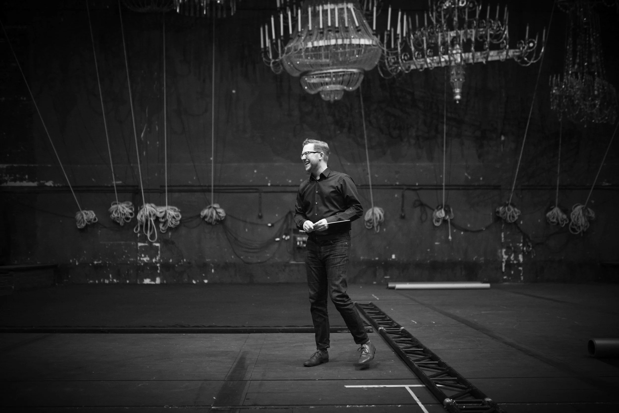 Black-and-white photo of Benjamin Bayl on theater stage beneath numerous ornate chandeliers