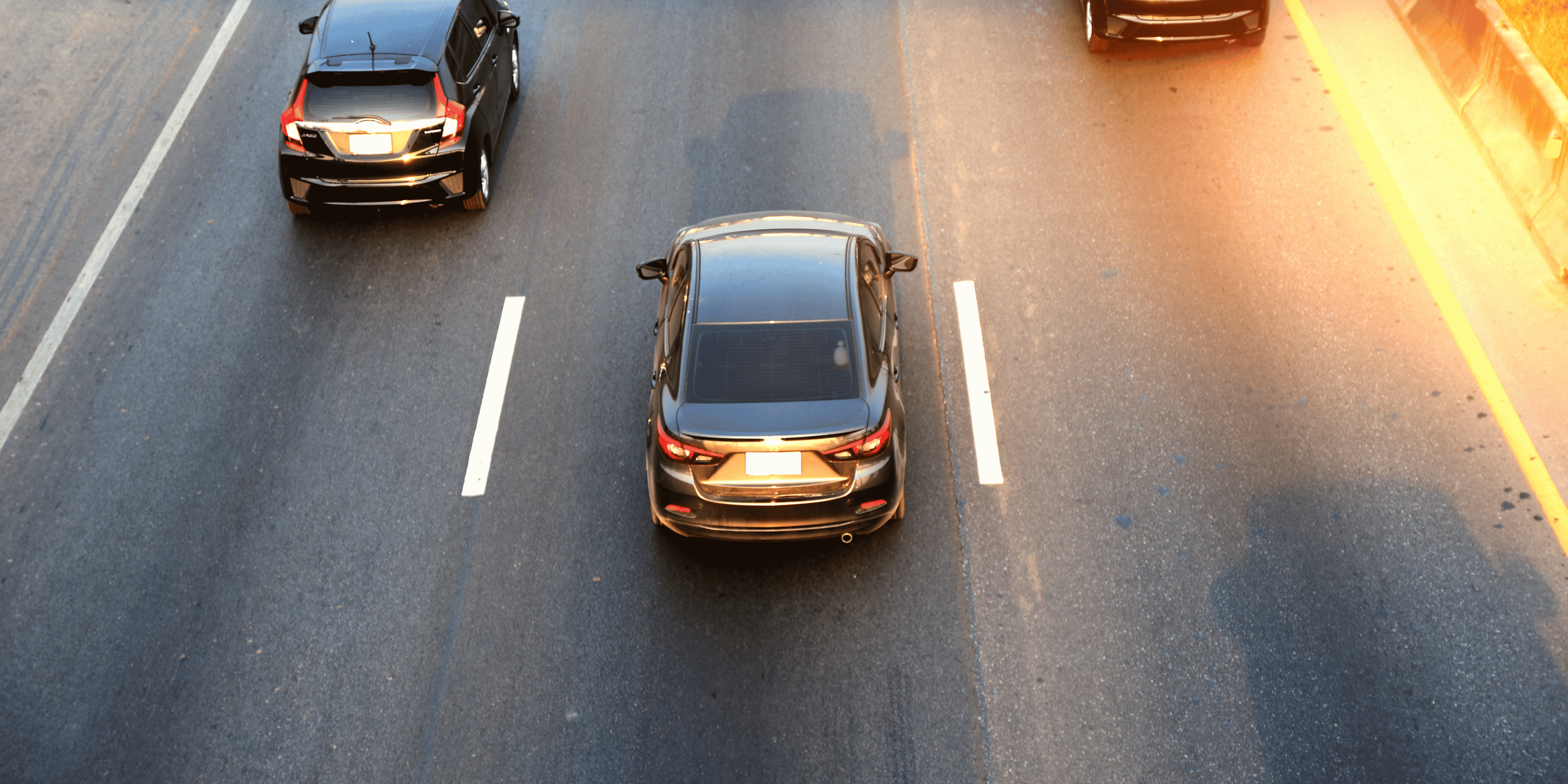 A top view of a parking lot with four black cars, three parked in designated spaces and one moving along the lane marked with white lines, with sunlight casting shadows and reflections on the cars.