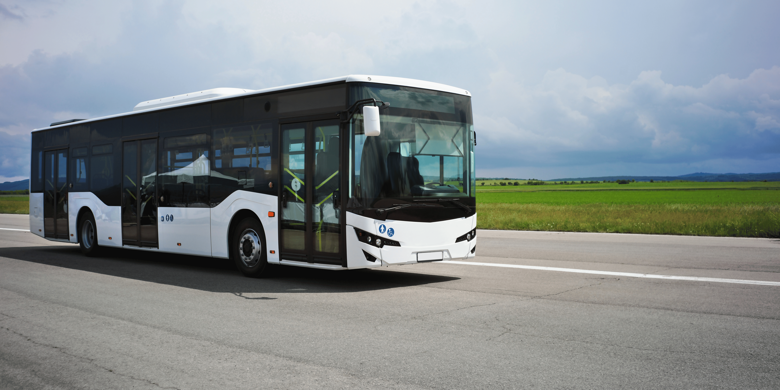 A modern white bus traveling on a highway through a rural landscape with green fields and mountains in the background under a partly cloudy sky.