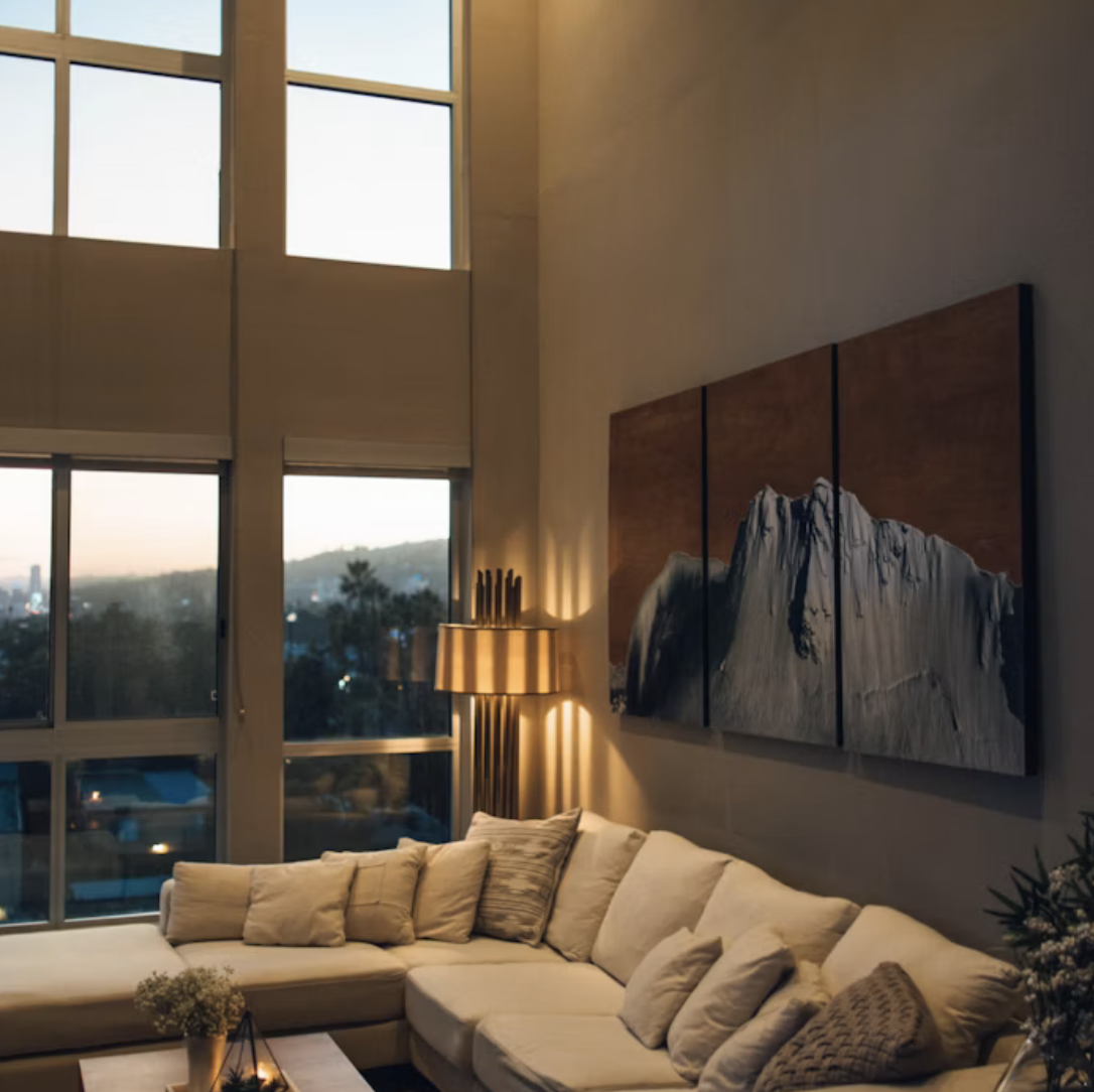 Interior of a modern living room with large windows, a beige sectional sofa with pillows, a wooden wall art piece, a floor lamp, and a view of the evening cityscape outside.