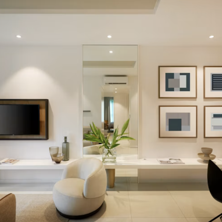 Modern living room with a wall-mounted TV, a white armchair, a glass vase with green leaves on a long white console, and a mirror reflecting part of the room. Artistic framed prints are on the wall.