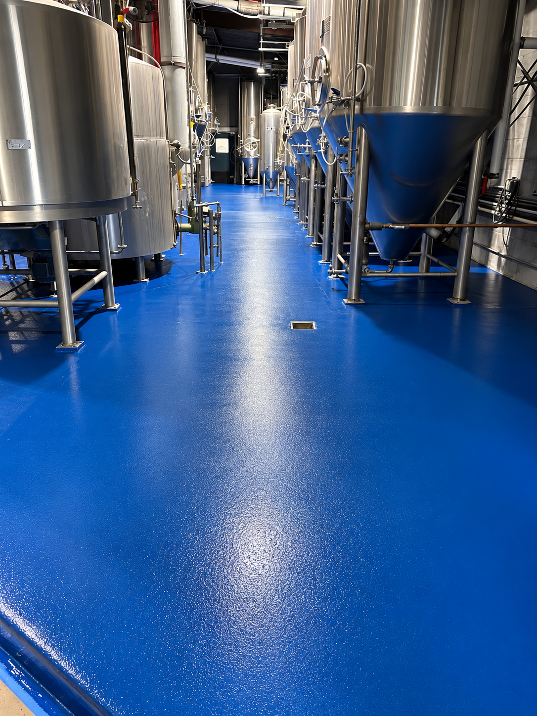 Industrial brewery equipment with steel tanks and pipes on a bright blue coated floor.