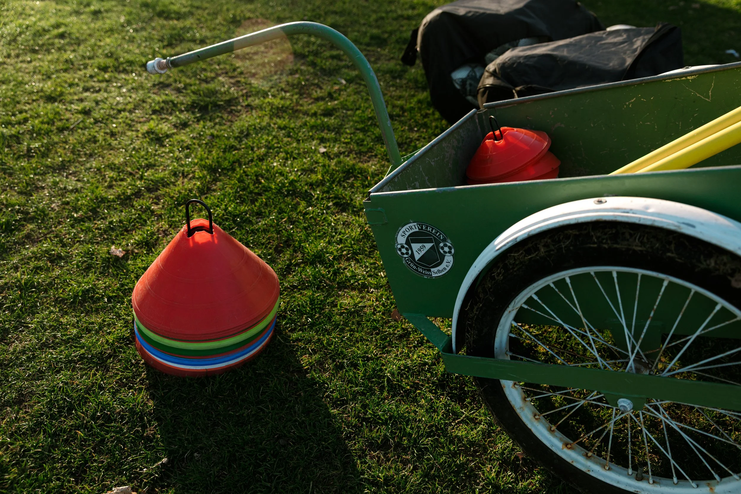 Eine grüne Handwagen mit einem Logo, auf dem das Jahr 1959 und Fußballsymbole stehen, daneben ein Stapel farbiger Fähnchen auf grünem Rasen.
