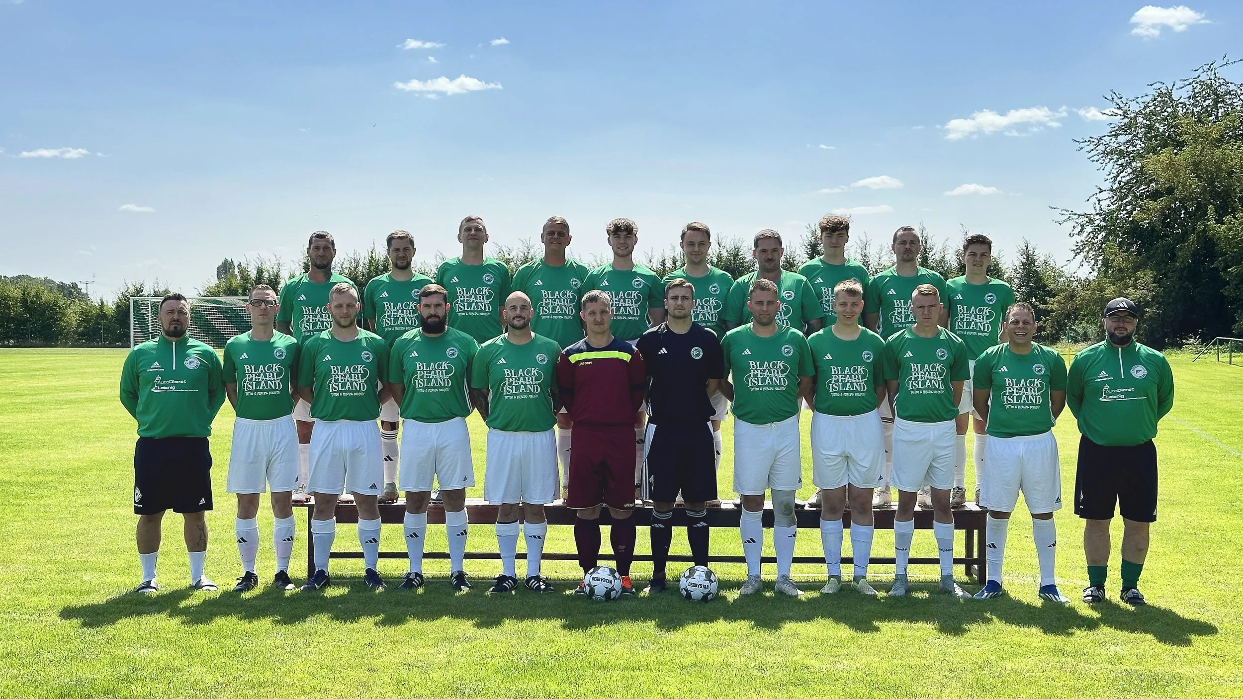 Mannschaftsfoto eines Fußballteams im Freien auf dem Spielfeld, bestehend aus 16 männlichen Spielern und zwei Trainern, alle in grünen Trikots mit Slogan "Black Pearl Island" und weißen Shorts, umgeben von Bäumen und einem blauen Himmel.