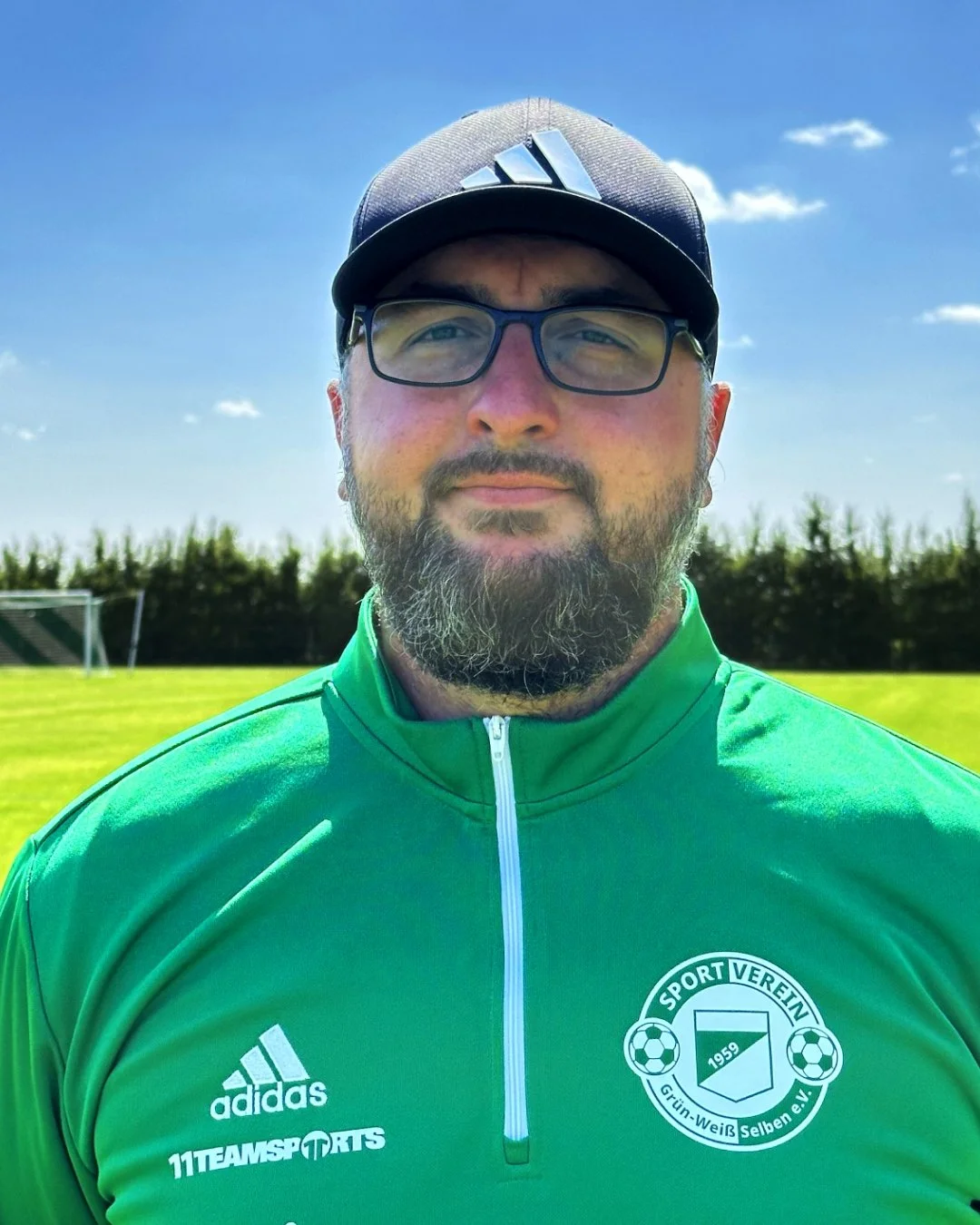 Mann mit Brille, schwarzem Cap und grüner Sportjacke, standing auf einem Fußballplatz mit grünem Rasen und Tor im Hintergrund, bei sonnigem Himmel.