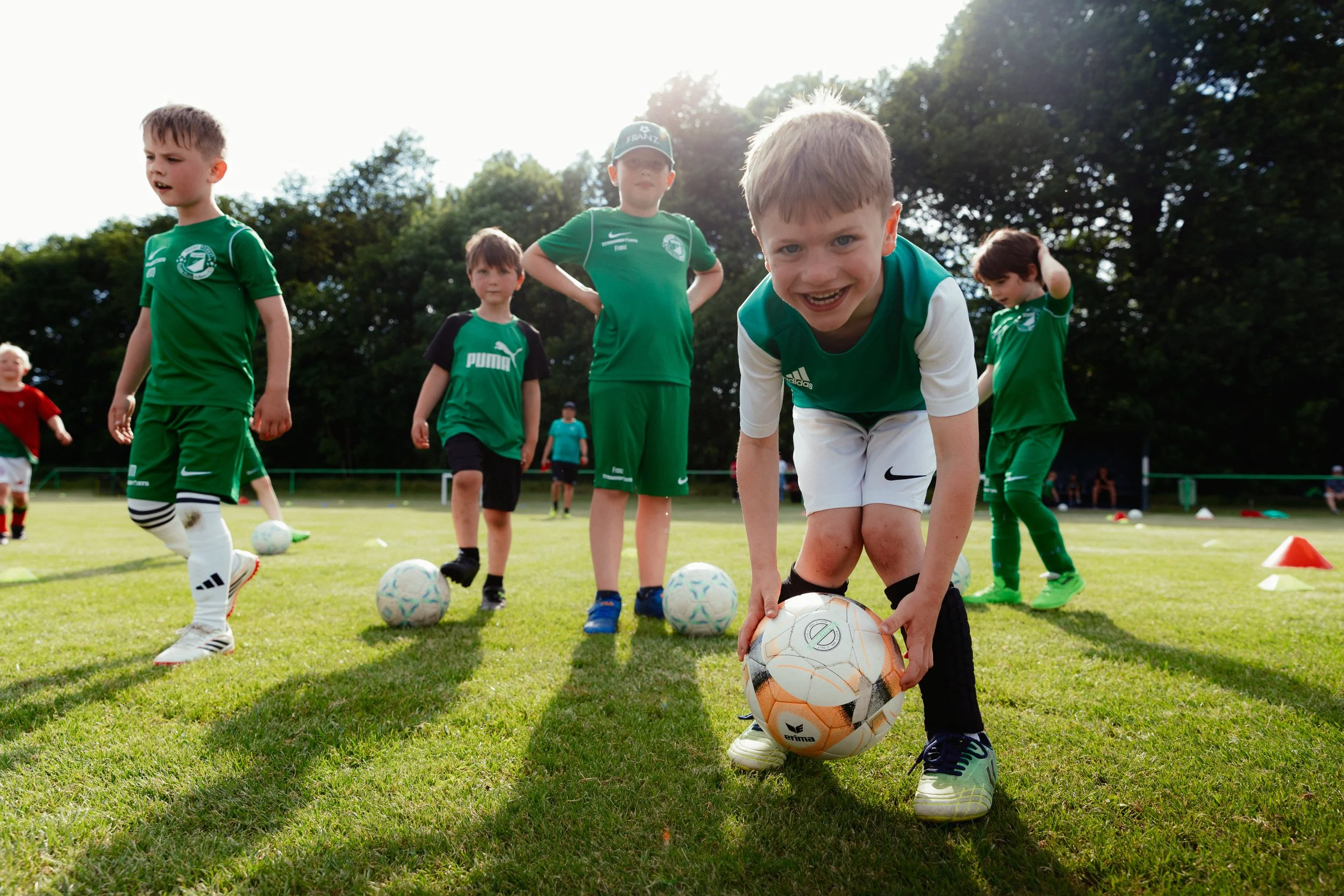 Junge Kinder beim Fußballtraining auf dem Rasen, mit bunten Bällen und Trainingsequipment, umgeben von Bäumen an einem sonnigen Tag