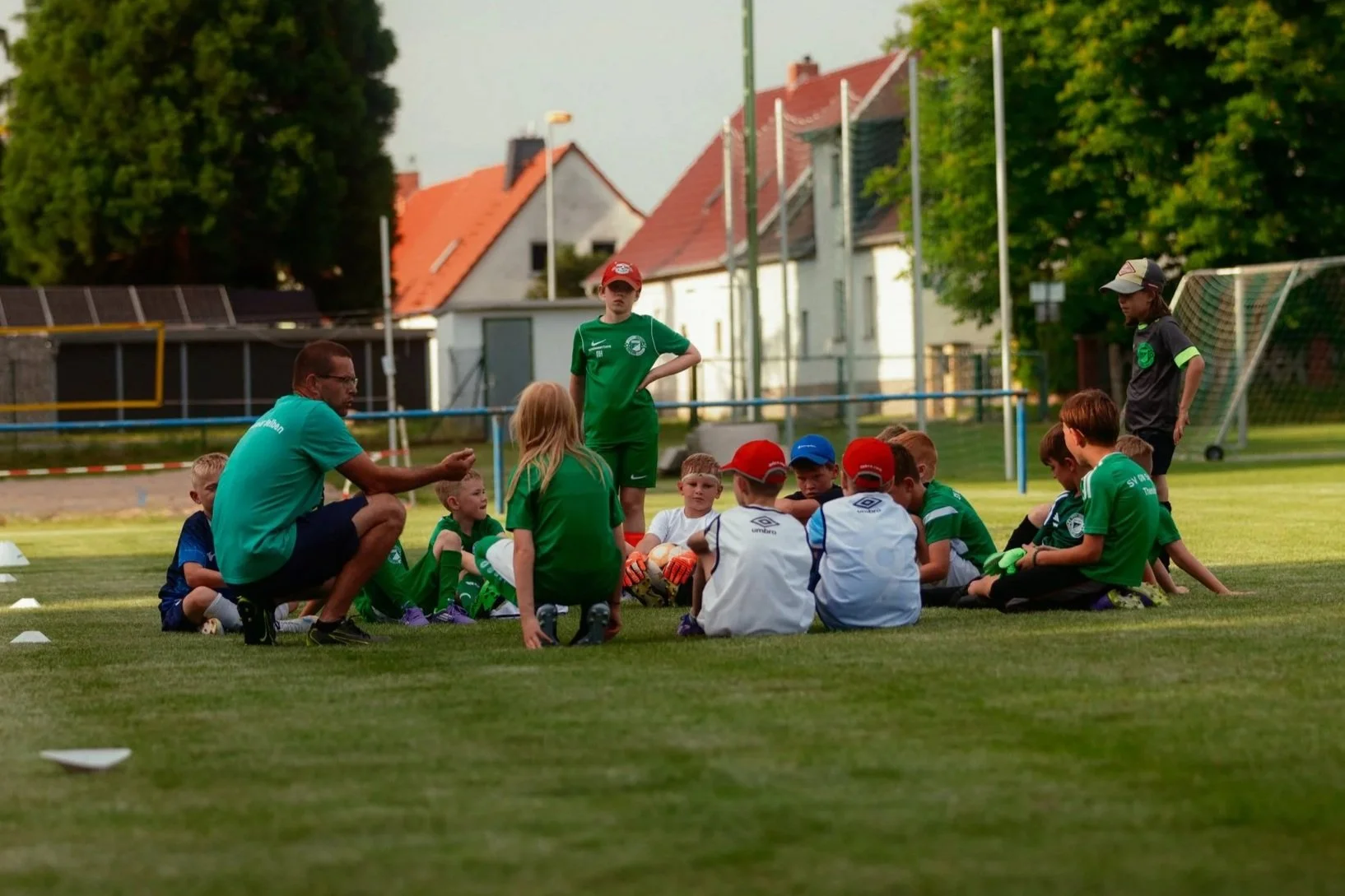 Kinder eines Fußballtrainings sitzen im Kreis auf dem Rasen, während ein Coach ihnen Anweisungen gibt. Zwei Jugendliche stehen daneben. Es ist ein sonniger Nachmittag, im Hintergrund sind Bäume und Häuser sichtbar.