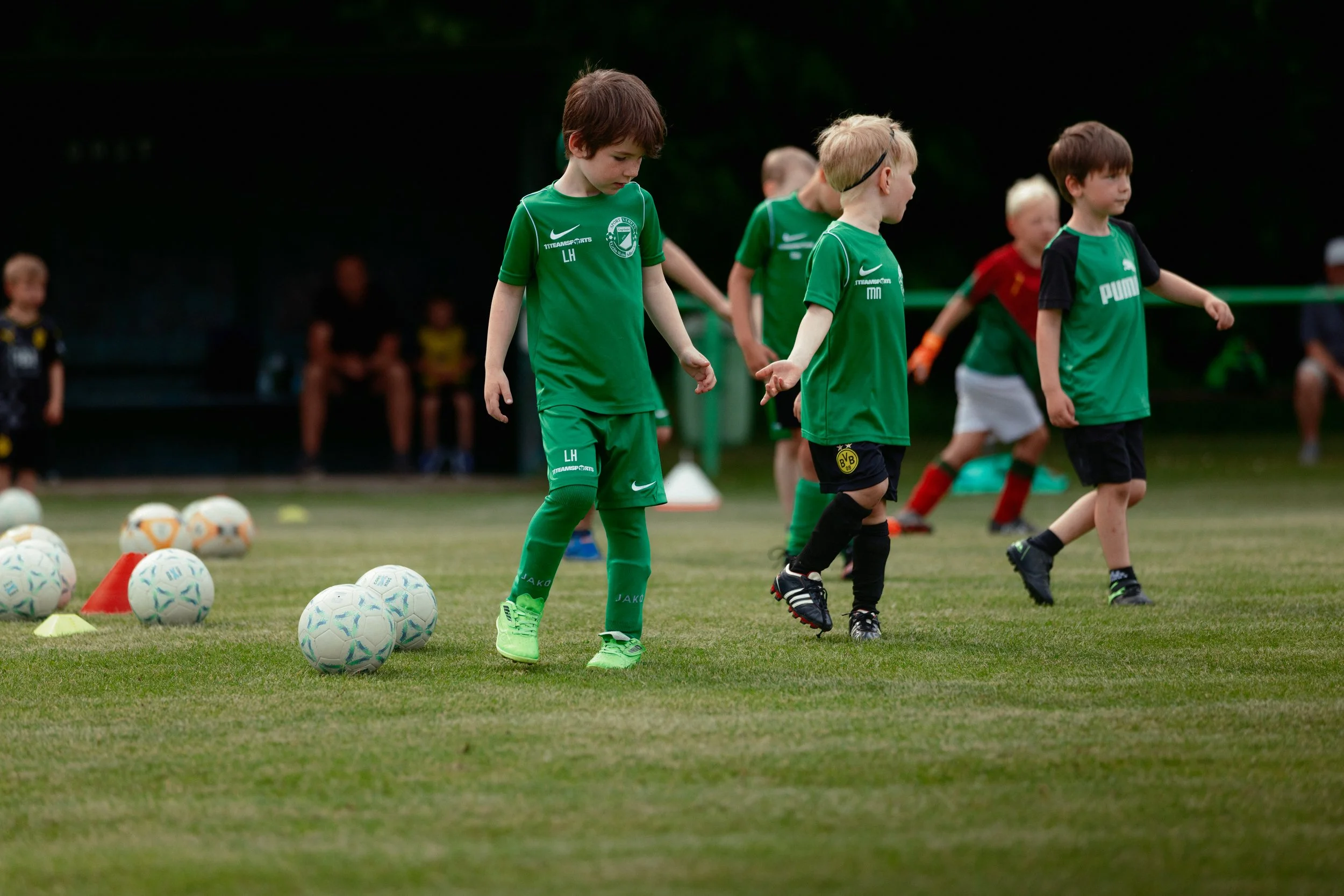 Junge Kinder beim Fußballtraining auf dem Platz, mit Fußballbällen und Markierungshütchen, in grünen Trikots.