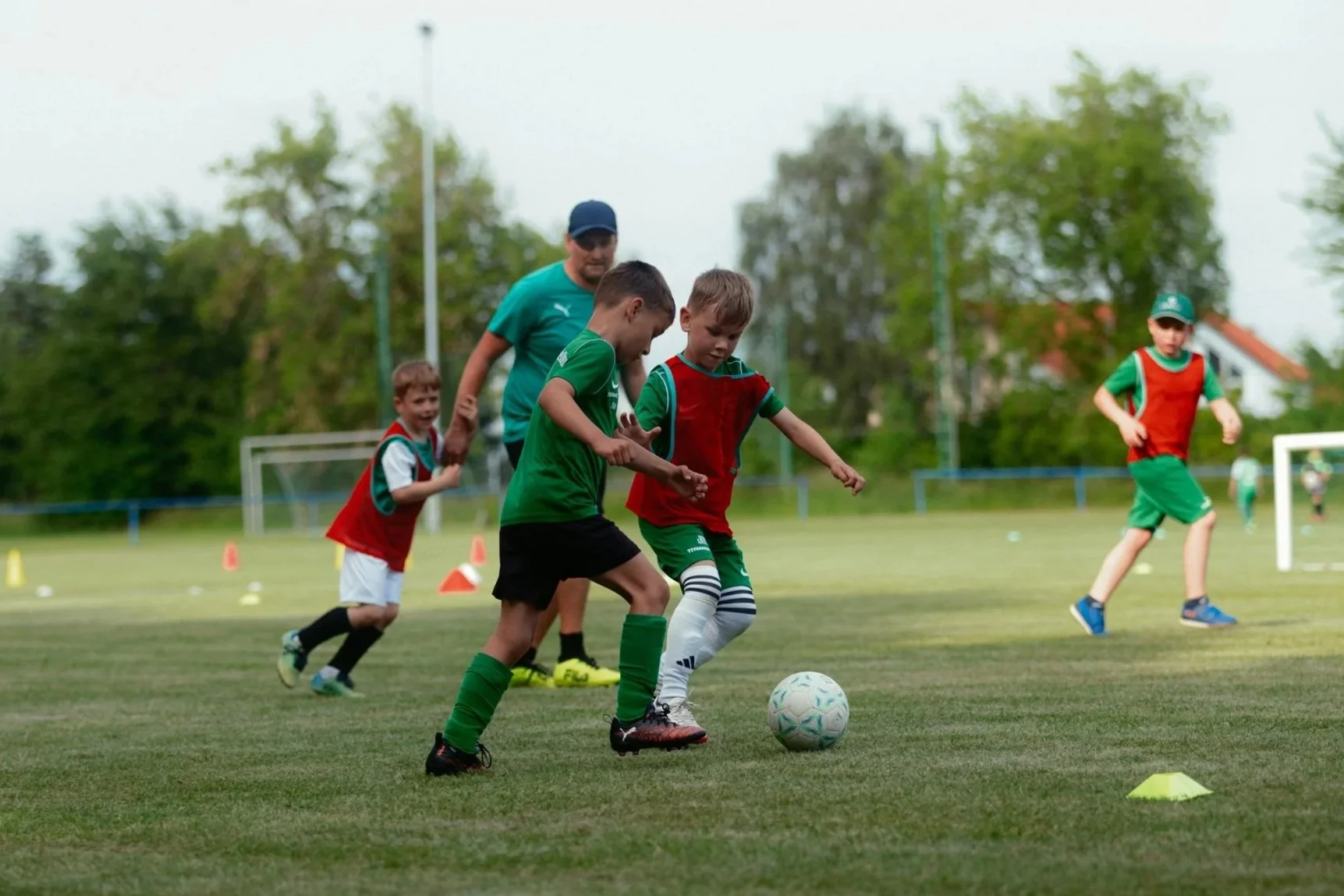 Junge Kinder spielen Fußball auf einem Sportplatz mit einem Erwachsenen im Hintergrund, alle tragen Sportkleidung und sind in Bewegung.
