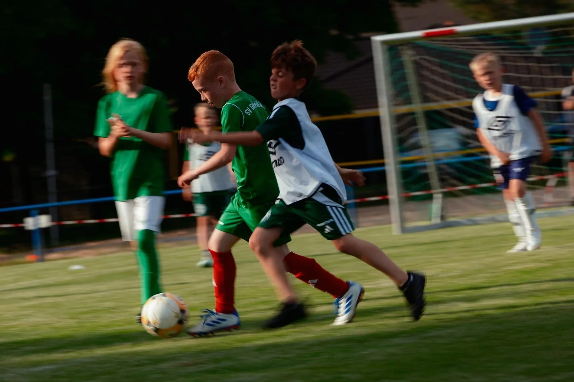 Jugendliche spielen Fußball auf dem Sportplatz, zwei Kinder raufen um den Ball, einer trägt ein grünes Trikot, ein anderer ein weißes Trikot mit schwarzen Details, im Hintergrund sind weitere Kinder und ein Tor sichtbar.