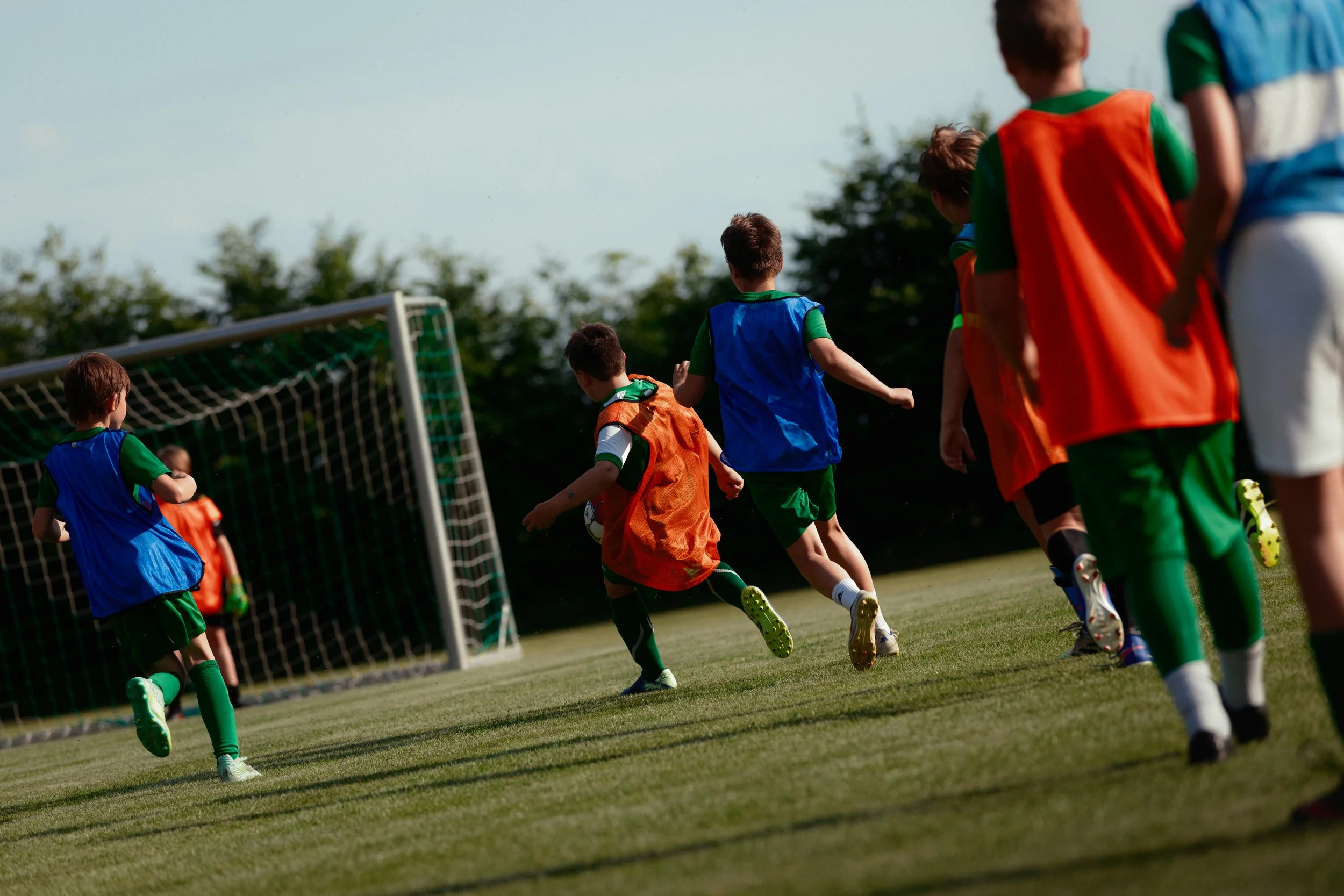 Junge Kinder spielen Fußball auf einem Sportplatz, tragen bunte Trikots und trainieren vor einem Tor.