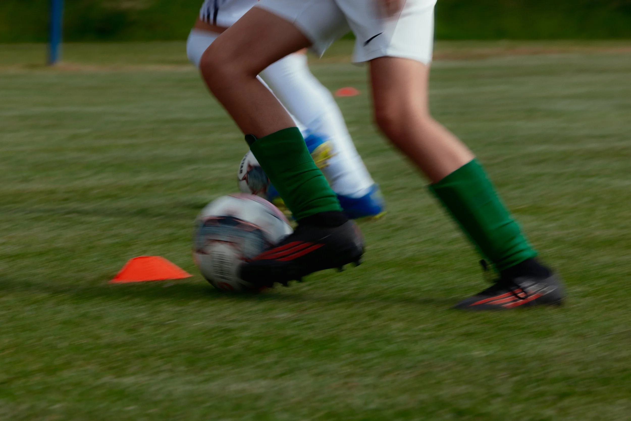 Fußballspieler beim Dribbling auf einem grünen Rasenplatz, abgekürzte Knie- und Beinregion, türkisch-grüne Stutzen, Fußball, orangehohes Hindernis, im Hintergrund Bäume