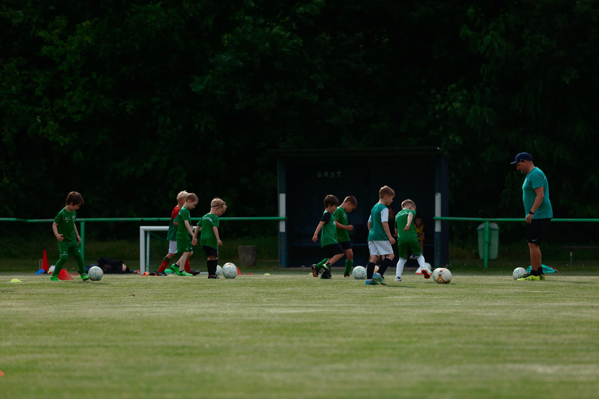 Jugendliche beim Fußballtraining auf dem Rasen, Trainer beobachtet, mit Bäumen im Hintergrund.