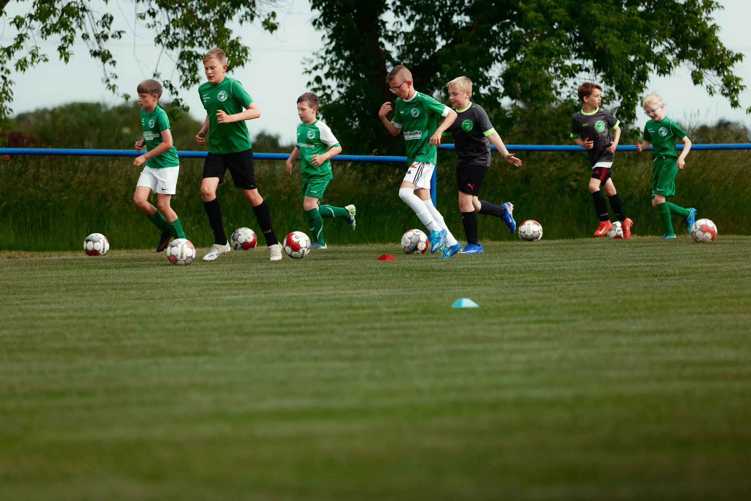 Gruppenspieler beim Fußballtraining auf einem grünen Rasenplatz, tragen grüne und schwarze Trikots, stehen in Linie neben Bällen und Markierungen, im Hintergrund Bäume.