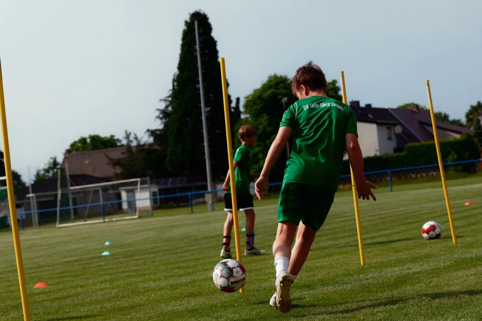 Zwei Kinder trainieren auf einem Fußballfeld, während eines von ihnen den Ball kontrolliert, umgeben von Hütchen, im Hintergrund Bäume und Häuser.
