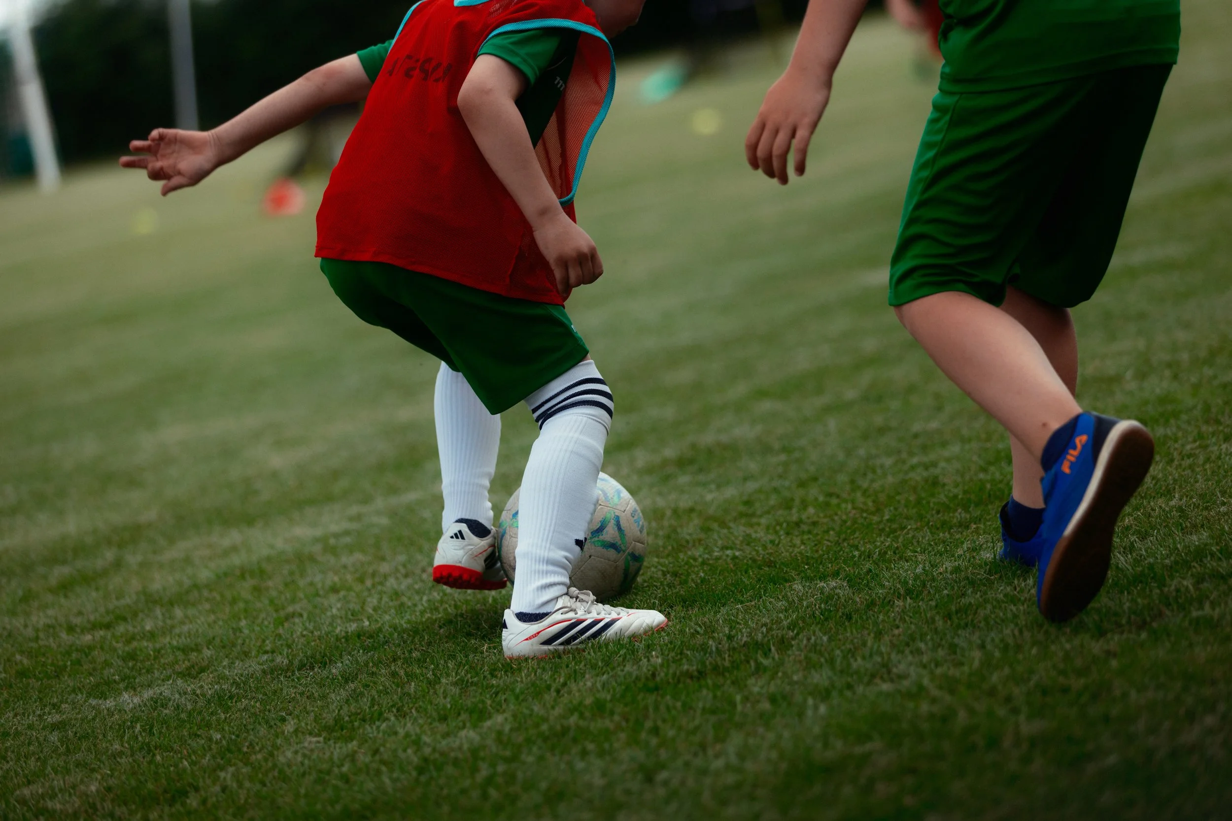 Ein Kind trägt ein rotes Fußballtrikot und weiße Stulpen, während es einen Ball auf einem grünen足球feld dribbelt, begleitet von einem Jugendlichen in grüner Kleidung.