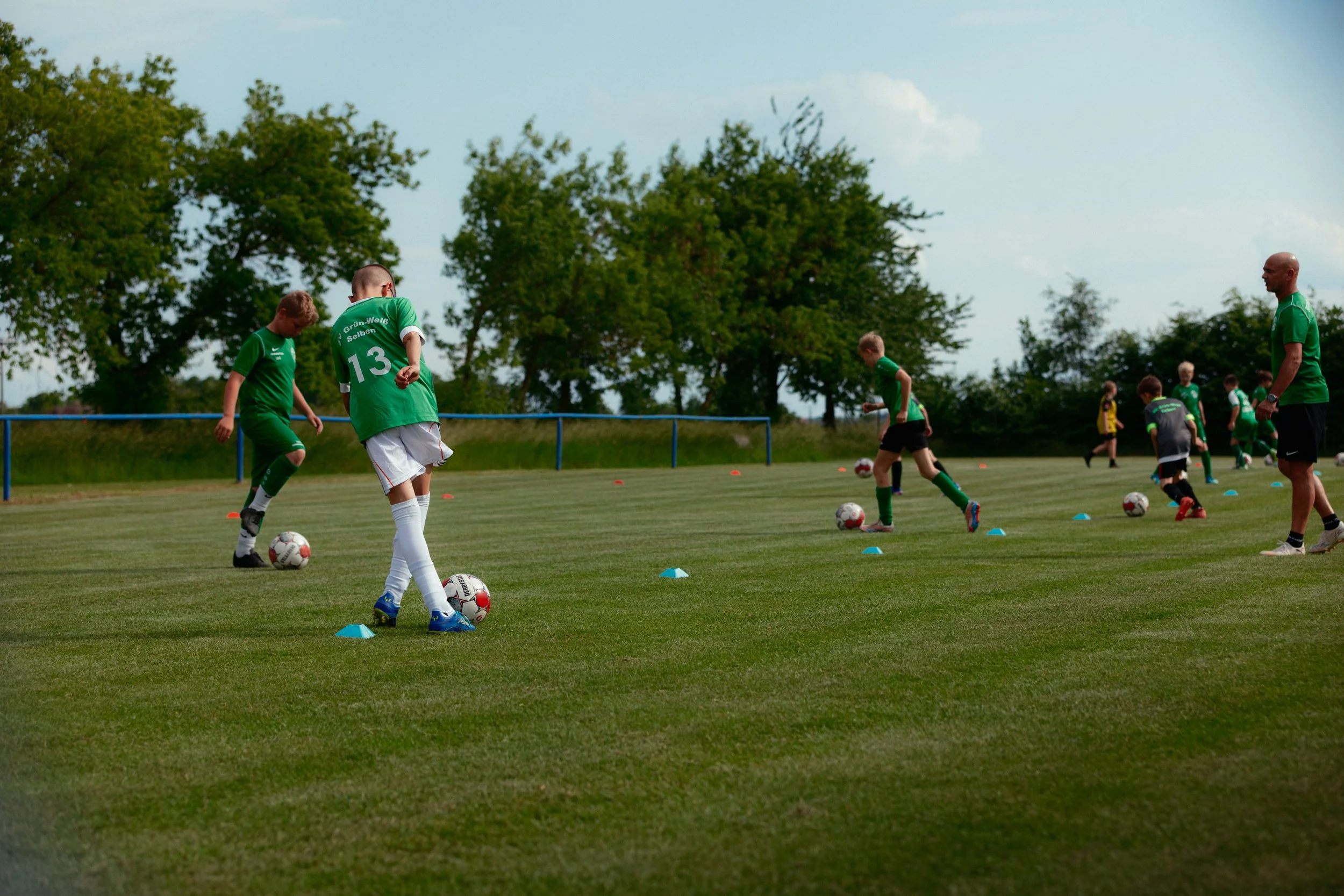 Jugendliche beim Fußballtraining auf dem Rasenplatz, viele Bälle und kleine Hütchen als Markierungen, Trainer beobachtet die Übungen.