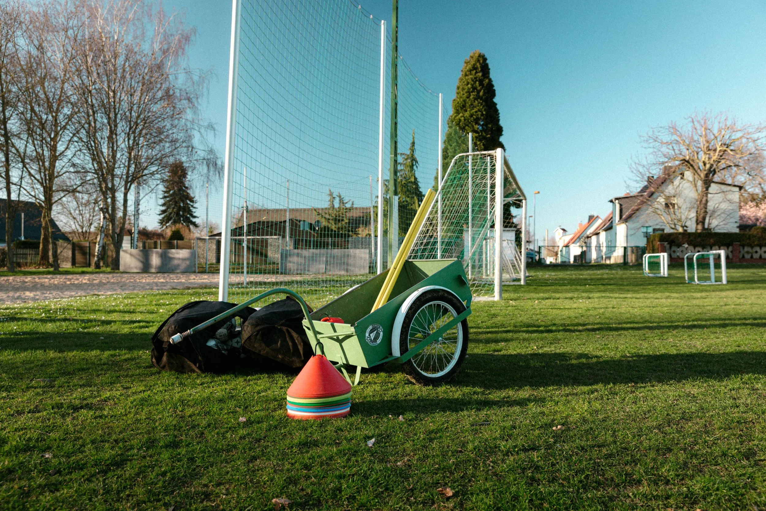 Ein fußballfeld mit Tor und Bälle, im Vordergrund eine grüne Sackkarre, sowie orange Verkehrshelme. Im Hintergrund sind Bäume und Häuser bei sonnigem Wetter zu sehen.