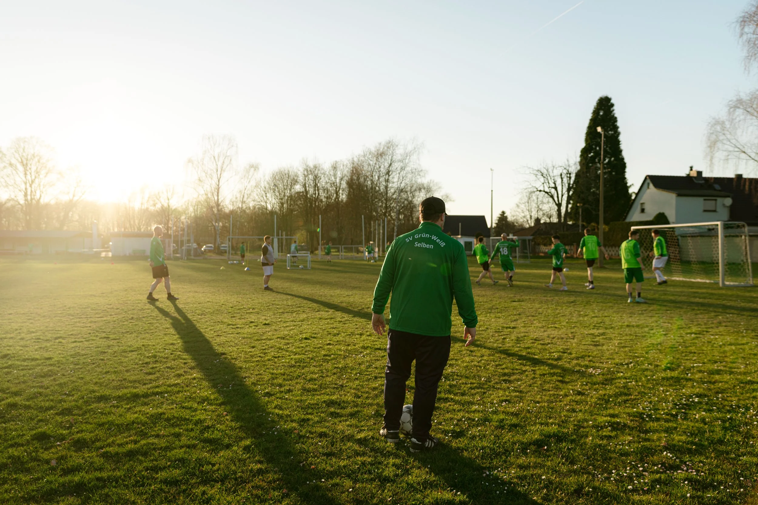 Fußballtraining auf dem Sportplatz bei Sonnenuntergang mit mehreren Spielern in grünen Trikots und einem Tor im Hintergrund.