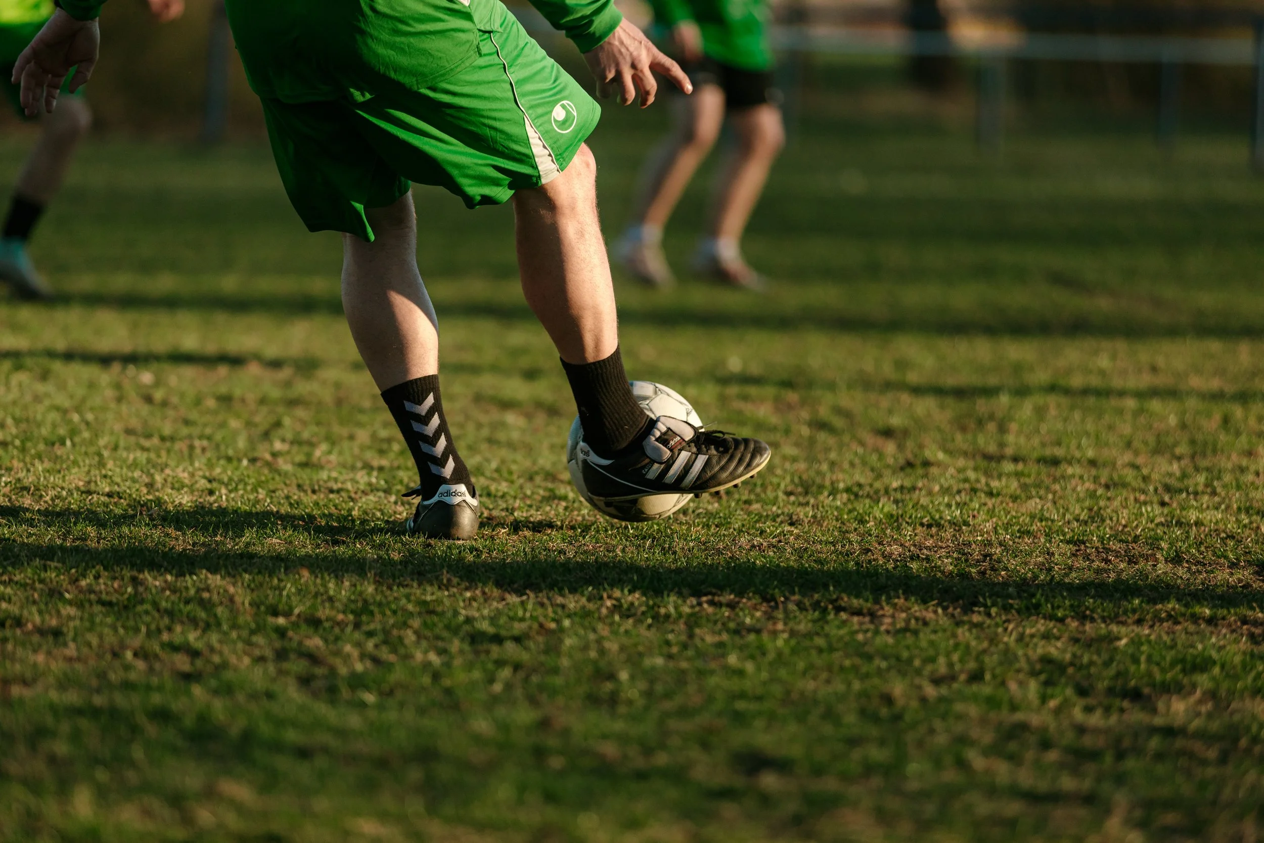 Fußballspieler in grüner Kleidung, der einen Ball auf einem Fußballfeld kickt, mit anderen Spielern im Hintergrund.
