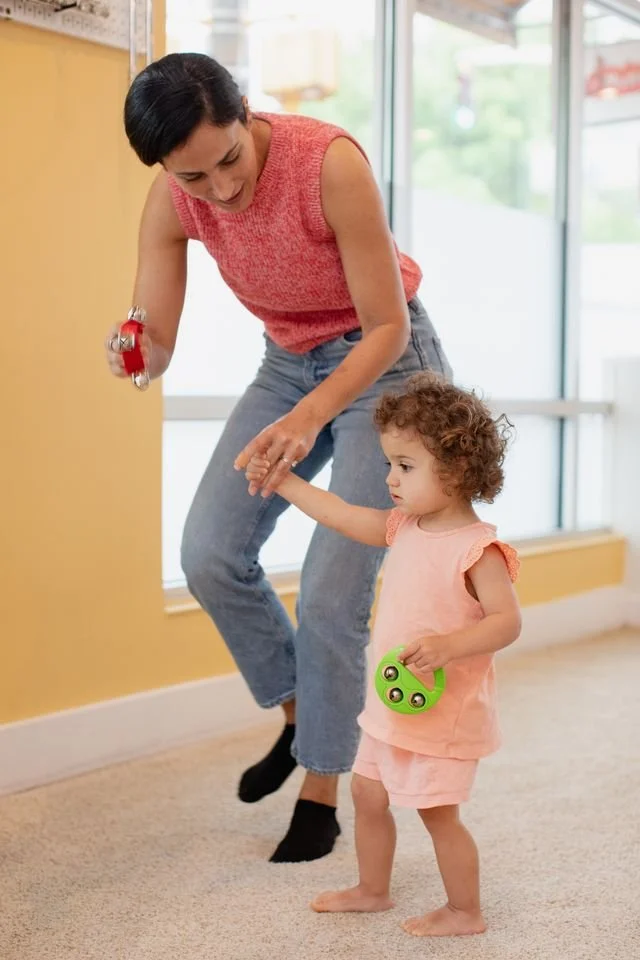 Madres y niuna habitación con paredes color amarillo y una ventana grande. La mujer está sonriendo y sosteniendo un objeto rojo en la mano, mientras la niña, con cabello rizado y vestida con un conjunto rosa, sostiene un cable de juguete color verde.