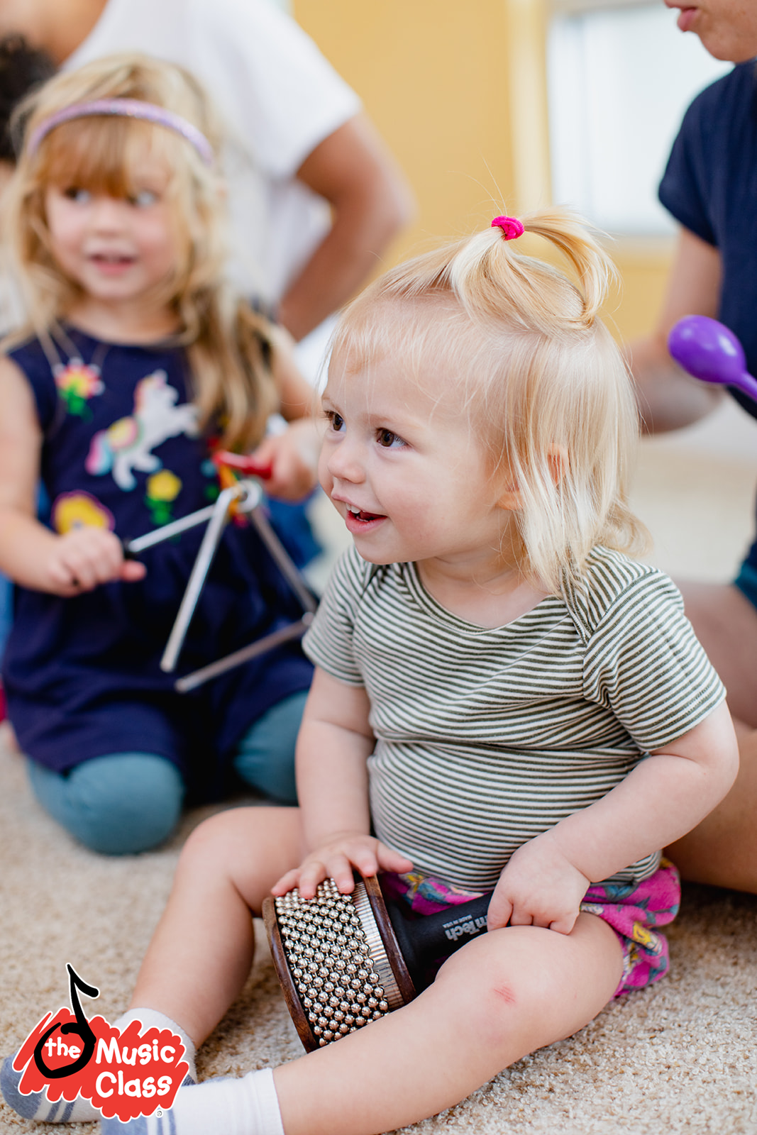 Niña rubia con camiseta de rayas jugando con un tambor en una clase de música, rodeada de otros niños.