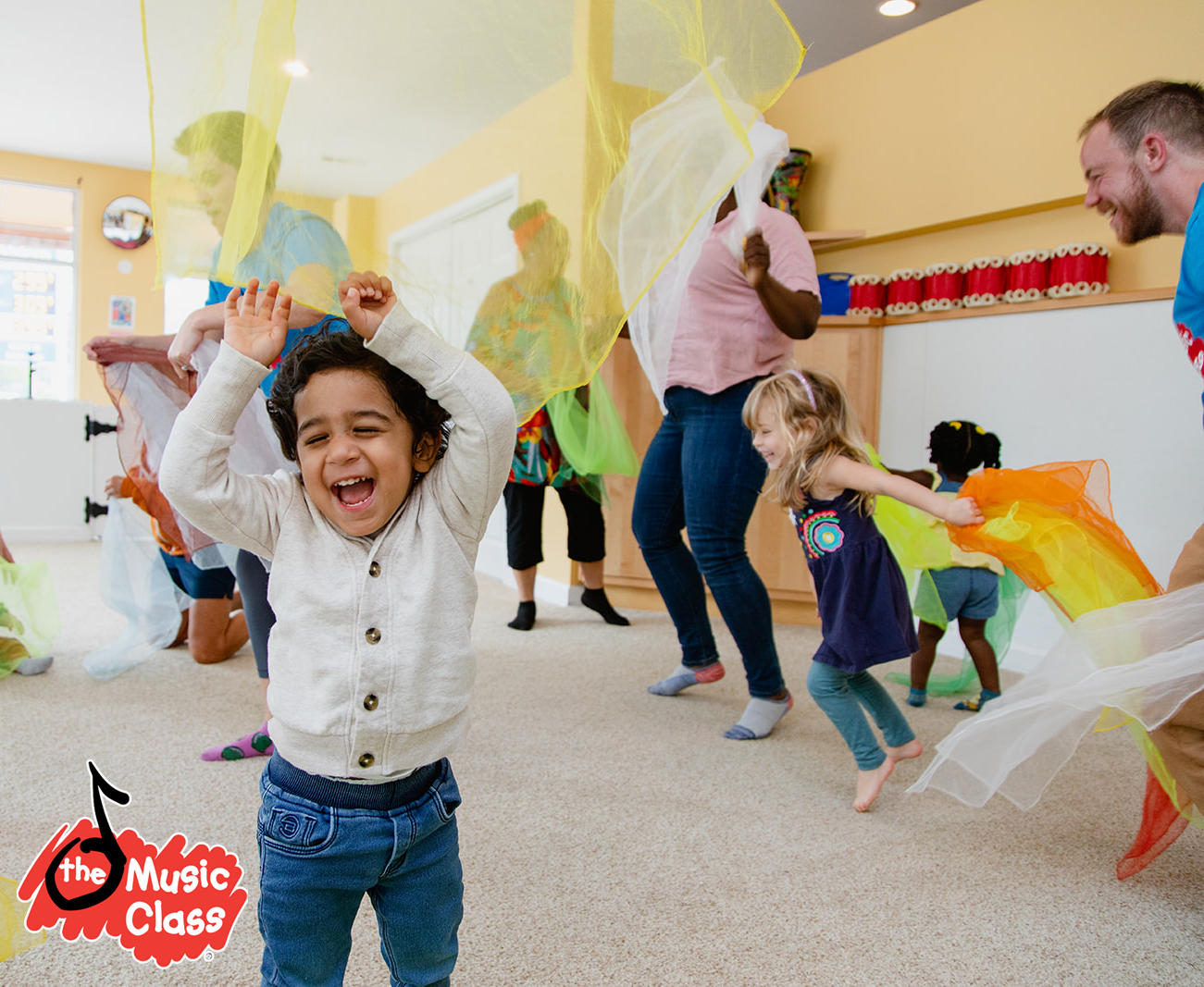 Niños y adultos bailando y jugando con telas de colores en una clase de música en un salón de clases.