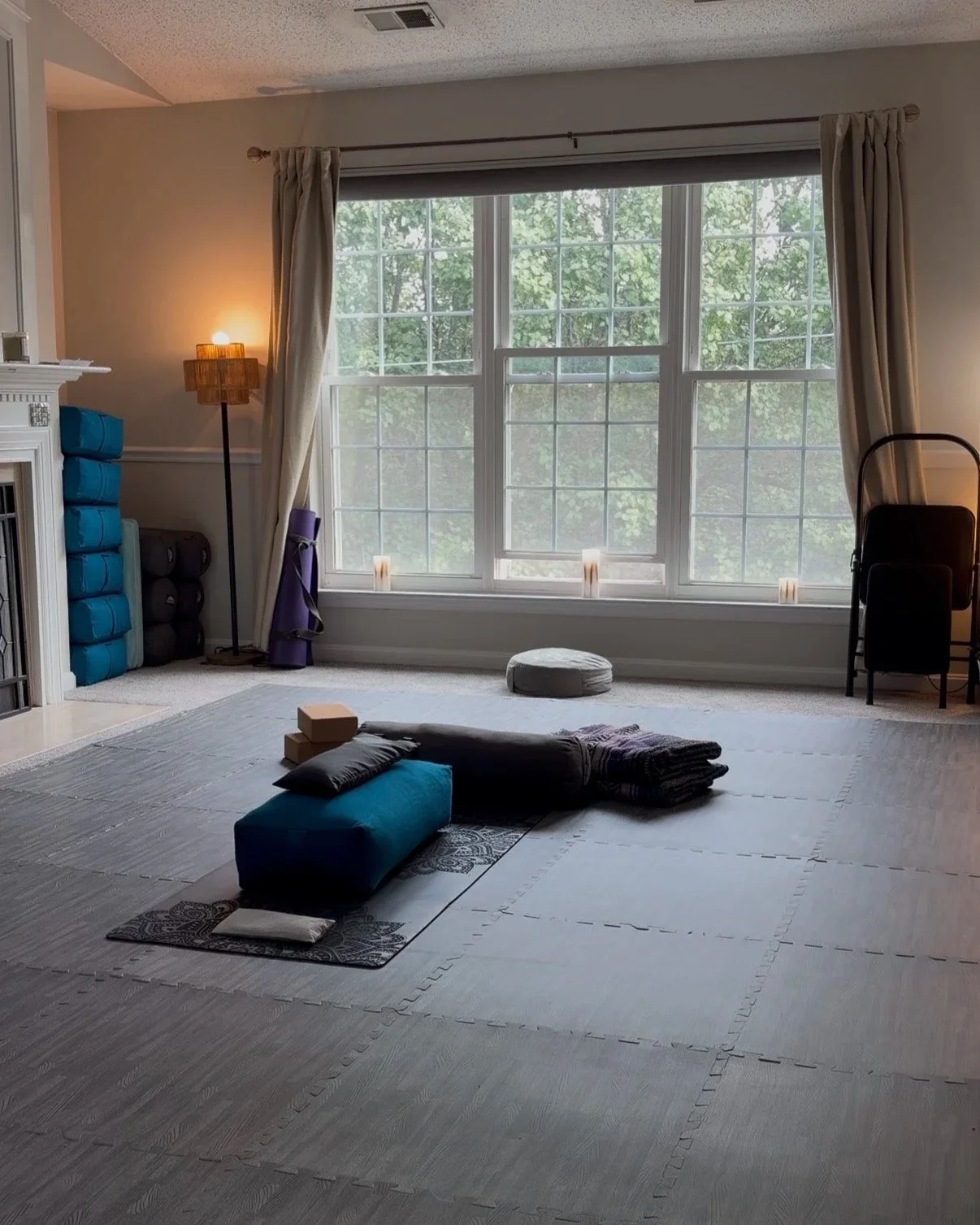 A room set up for yoga or meditation with a large window, yoga mats, blocks, a cushion, a folded blanket, candles, and a small round cushion, with a cozy and calming atmosphere.