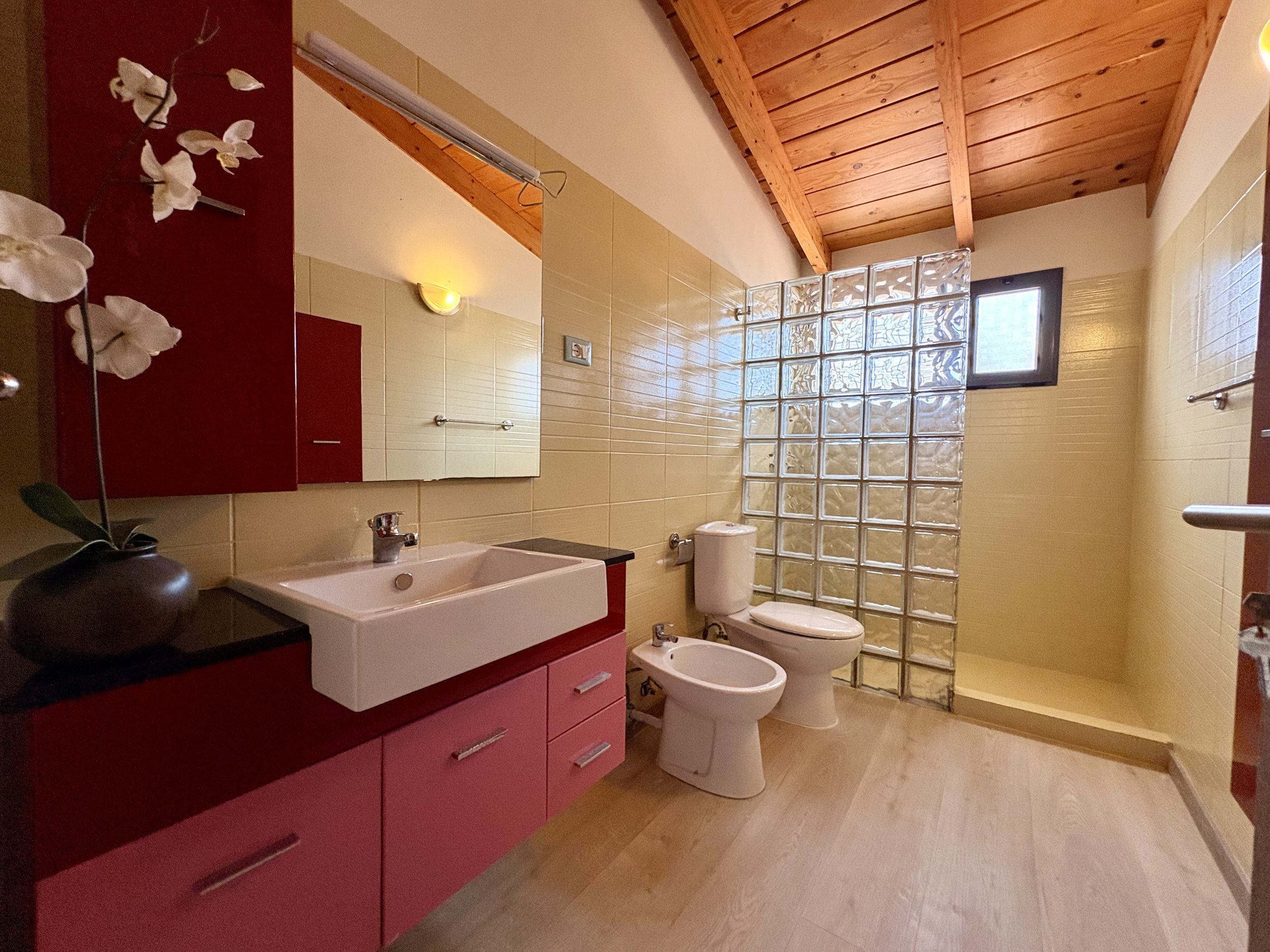 A bathroom with a red vanity cabinet and a white sink, beige tiled walls, a toilet, a bidet, a wall-mounted mirror, a towel rail, a window, a glass brick partition, and ceiling with wooden beams.