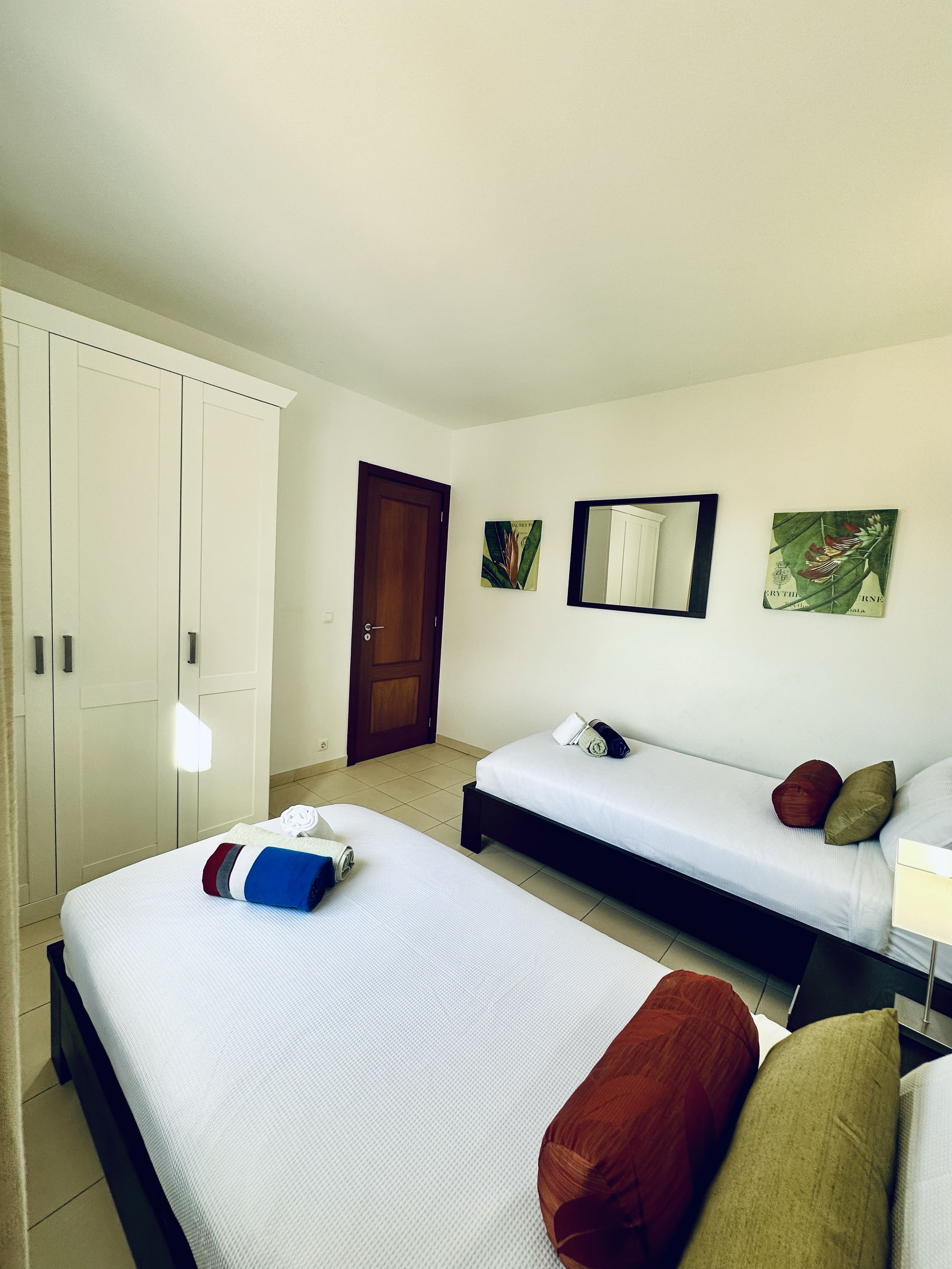 A hotel room with two single beds, pillows, and rolled towels, a wardrobe, a door, a mirror, and framed artwork on the wall.