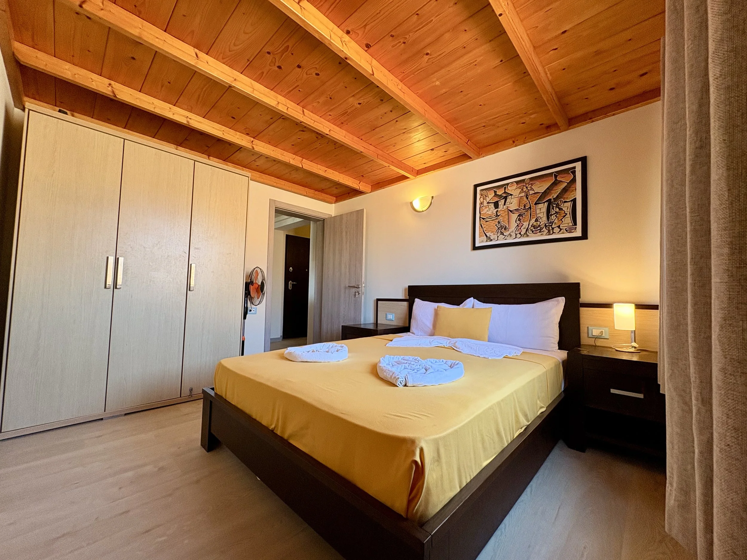 A bedroom with a wooden ceiling, a bed with yellow bedding, a nightstand with a lamp, a wardrobe, and a framed artwork on the wall.