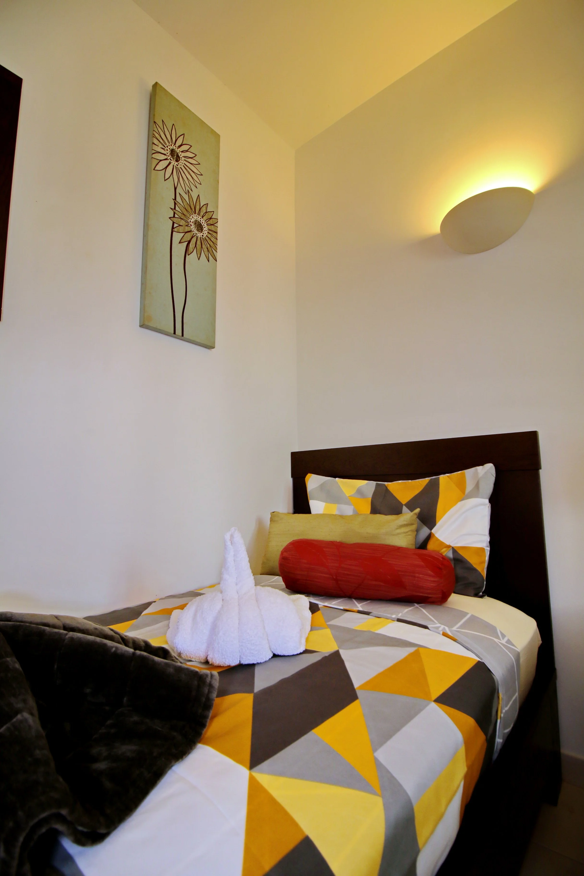 A bedroom with a bed that has a colorful geometric patterned bedspread in yellow, gray, and white. On the bed, there are folded white towels shaped like a swan, and a long red pillow. The headboard is dark wood, and there are multiple pillows, includ