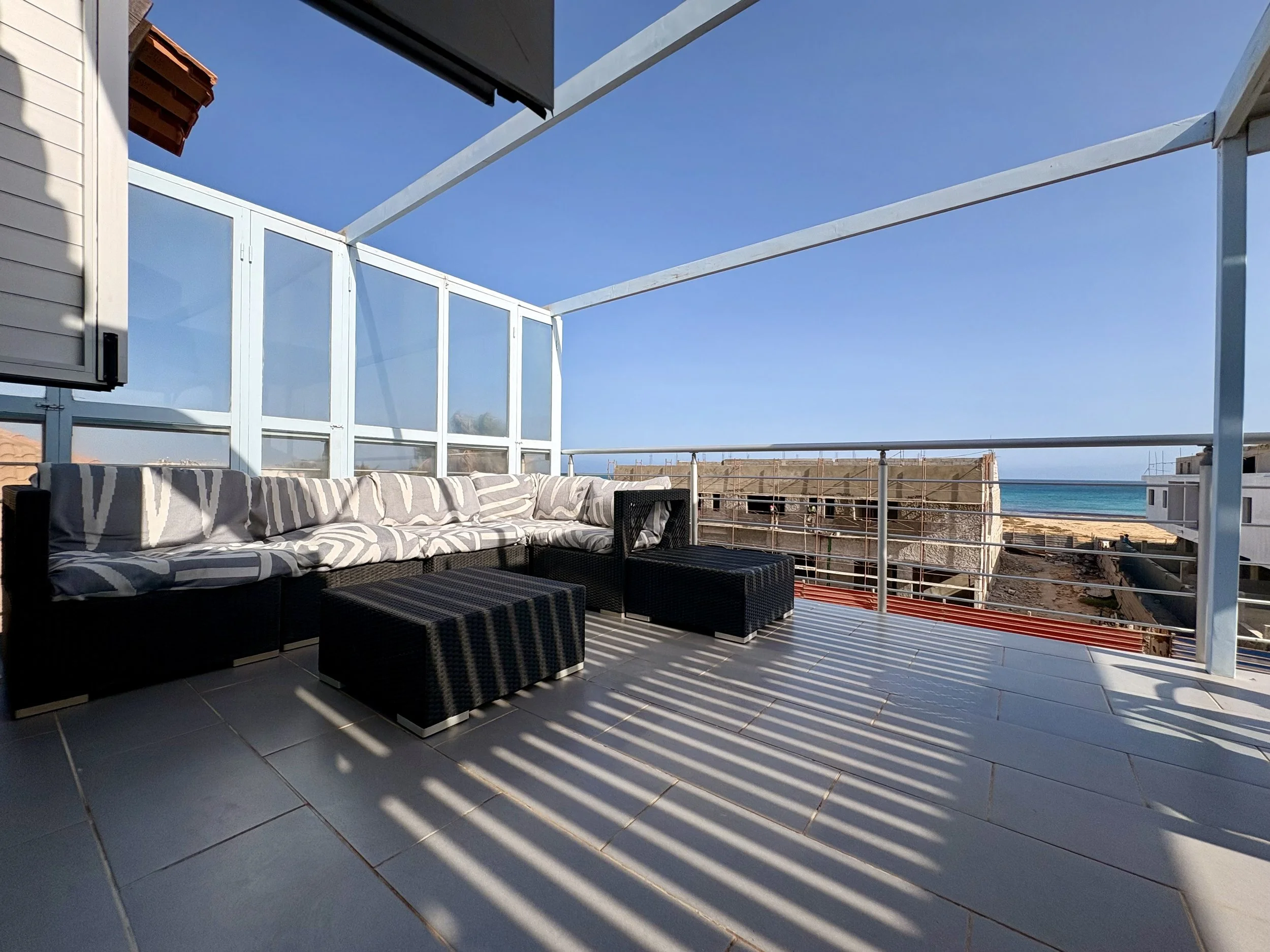 View of a modern balcony with striped outdoor furniture and glass panels, overlooking a beach and blue sky.