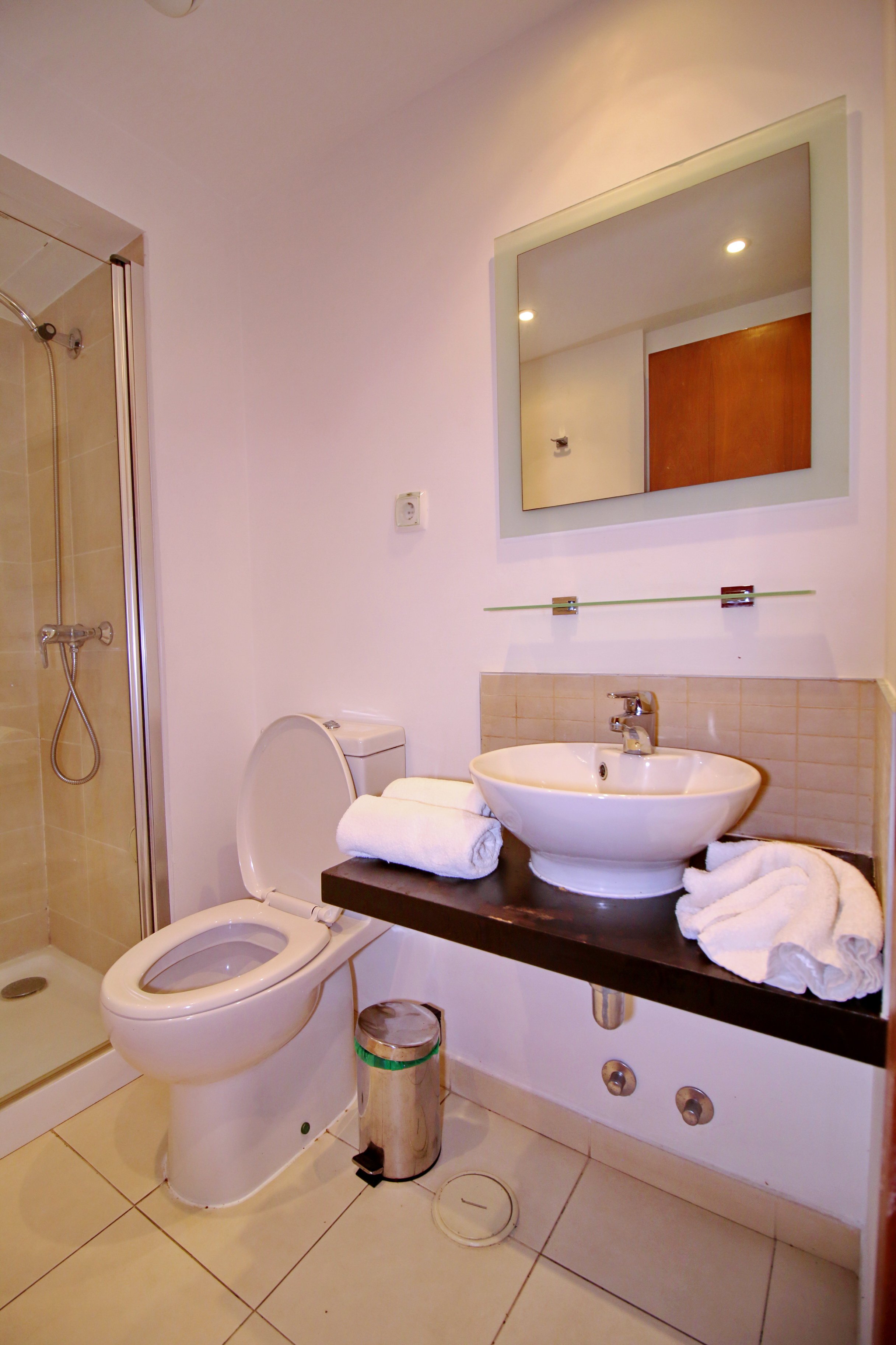 A bathroom with a shower, toilet, and a vanity with a vessel sink. Towels are rolled and placed on the vanity. There is a mirror above the sink and a trash can next to the toilet.