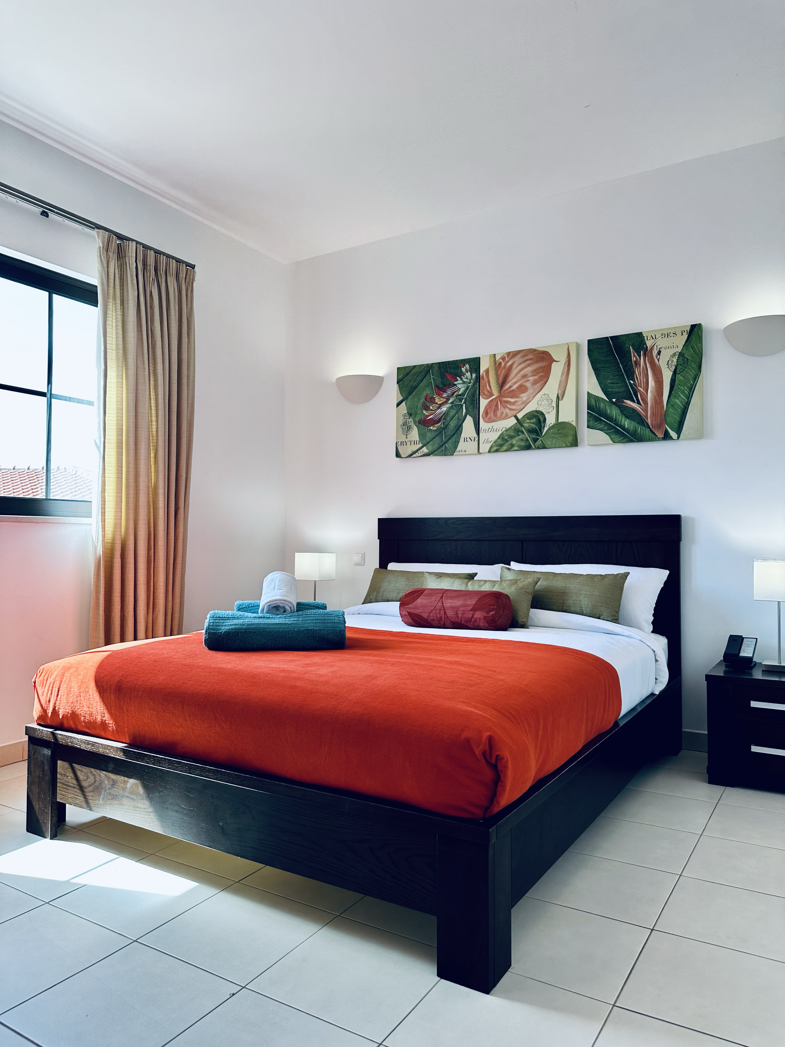 A modern bedroom with a large bed covered in a red blanket, green and red pillows, and a towel roll at the foot. The room has a window with beige curtains, wall-mounted lights, framed floral artwork above the bed, white walls, and tiled floors.