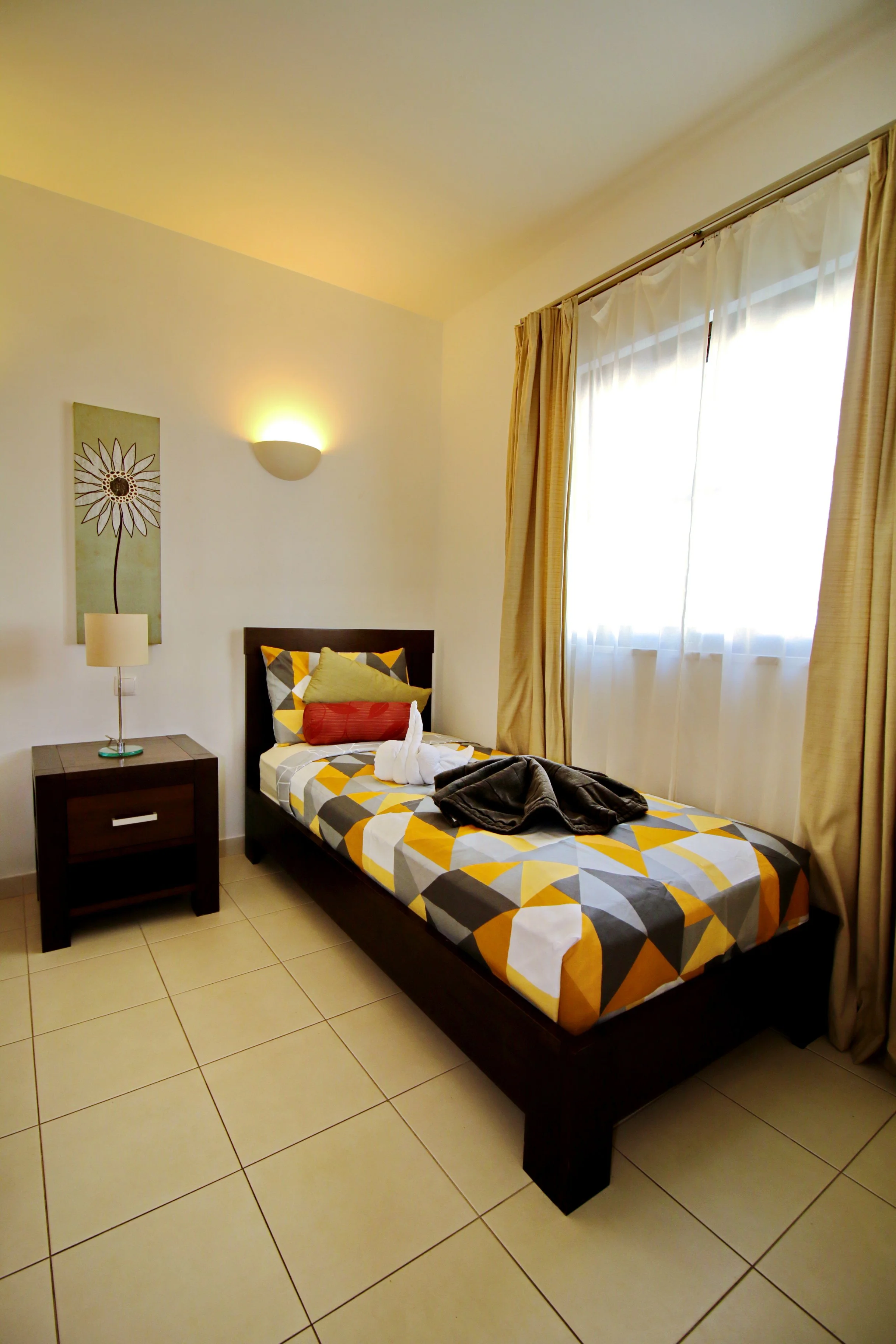 A single bed with a colorful geometric patterned bedspread, pillows, and towels folded on top, located in a bright room with white walls, a window with cream curtains, a wall sconce, a painting of a flower, and a small black bedside table.